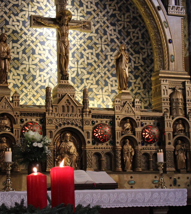 "Evangelischer Gottesdienst - Freude zu verschenken": Ein Adventskranz mit vier brennenden roten Kerzen steht im Vordergrund. Dahinter steht der Altar mit einem weißem Tischtuch, auf dem eine aufgeschlagene Bibel liegt. Darüber befindet sich ein aus Holz geschnitztes Altarbild mit Jesus am Kreuz mit einem Hintergrund aus blaubeigegemusterten Fliesen.