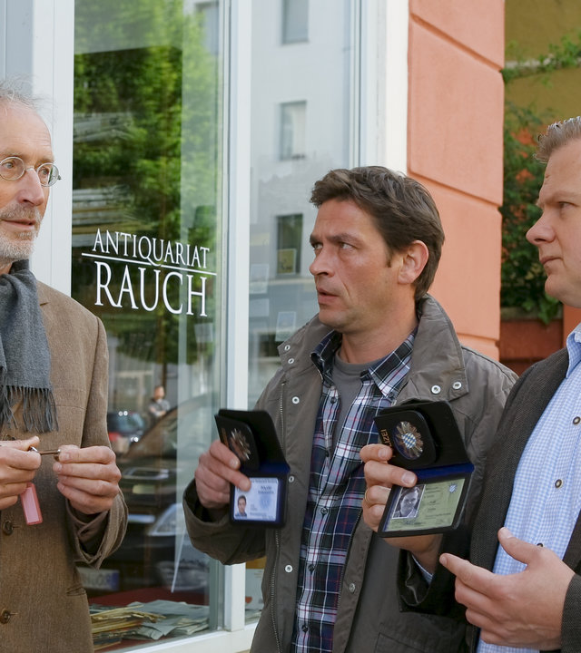 "Die Rosenheim-Cops: Jagd auf Watzmann": Vor dem Antiquariat befragen die Cops Lind (Tom Mikulla) und Hartl (Michael A. Grimm) Antiquar Anton Rauch (Frieder Venus). Sie halten ihm ihren Polizeiausweis hin.