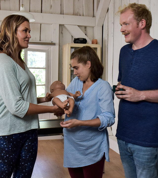 "Lena Lorenz - Baby auf Probe": In der Hebammenpraxis überreicht Lena Lorenz (Judith Hoersch) der schwangeren Alina Moosleitner (Anne-Marie Lux) eine Babypuppe. Daneben steht Tim Scheuer (Sebastian Griegel) und schaut freudig.