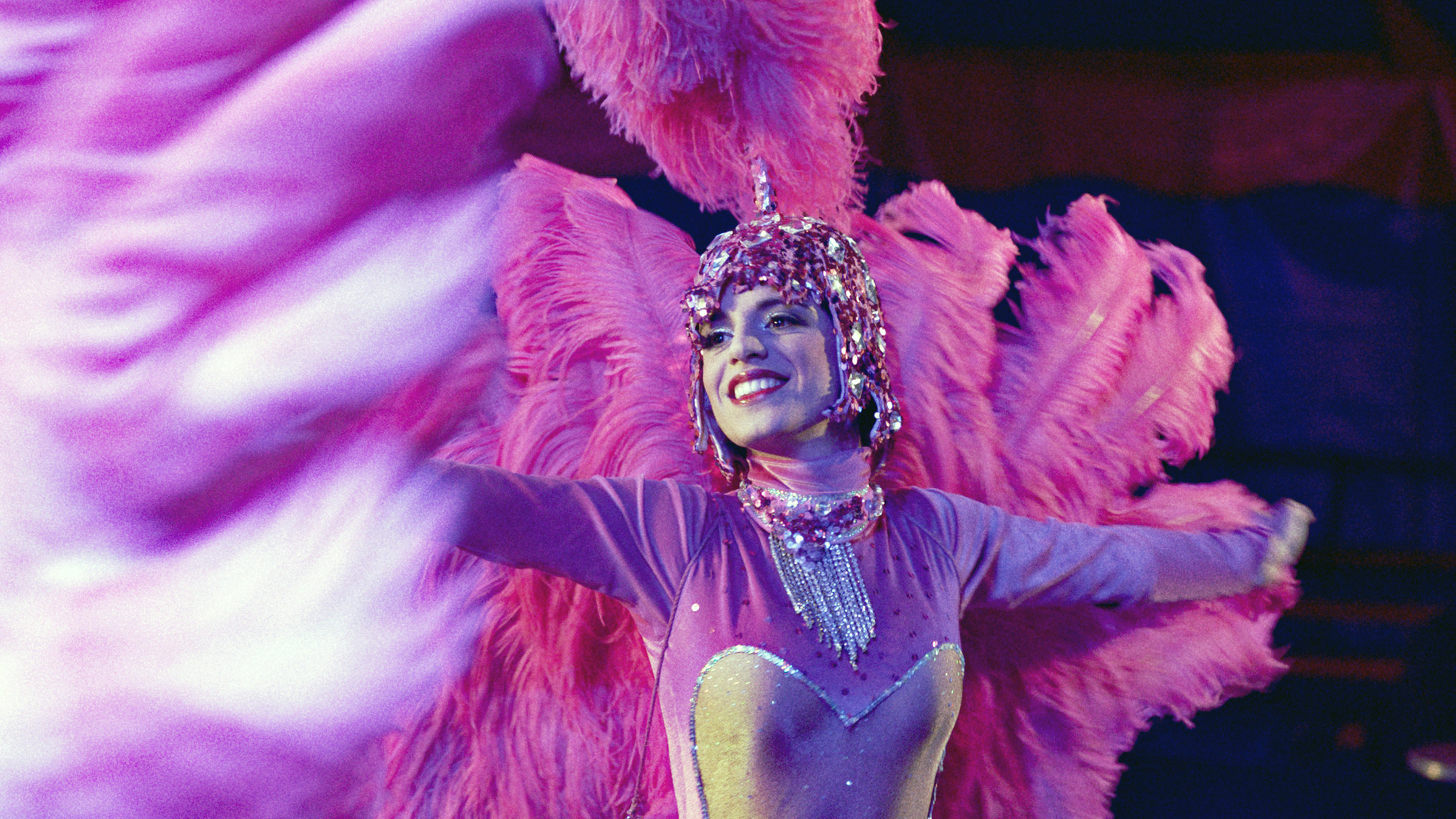 "KlassiXS: Macht und Hierarchien - Apollo und Artemisia": Eine Frau in einem rosa Tanzkostüm, mit engem Anzug, Kopfschmuck und Federn auf dem Rücken. Sie breitet die Arme aus und lächelt. Das Bild ist leicht verdeckt durch die Federn eines anderen Kostüms.