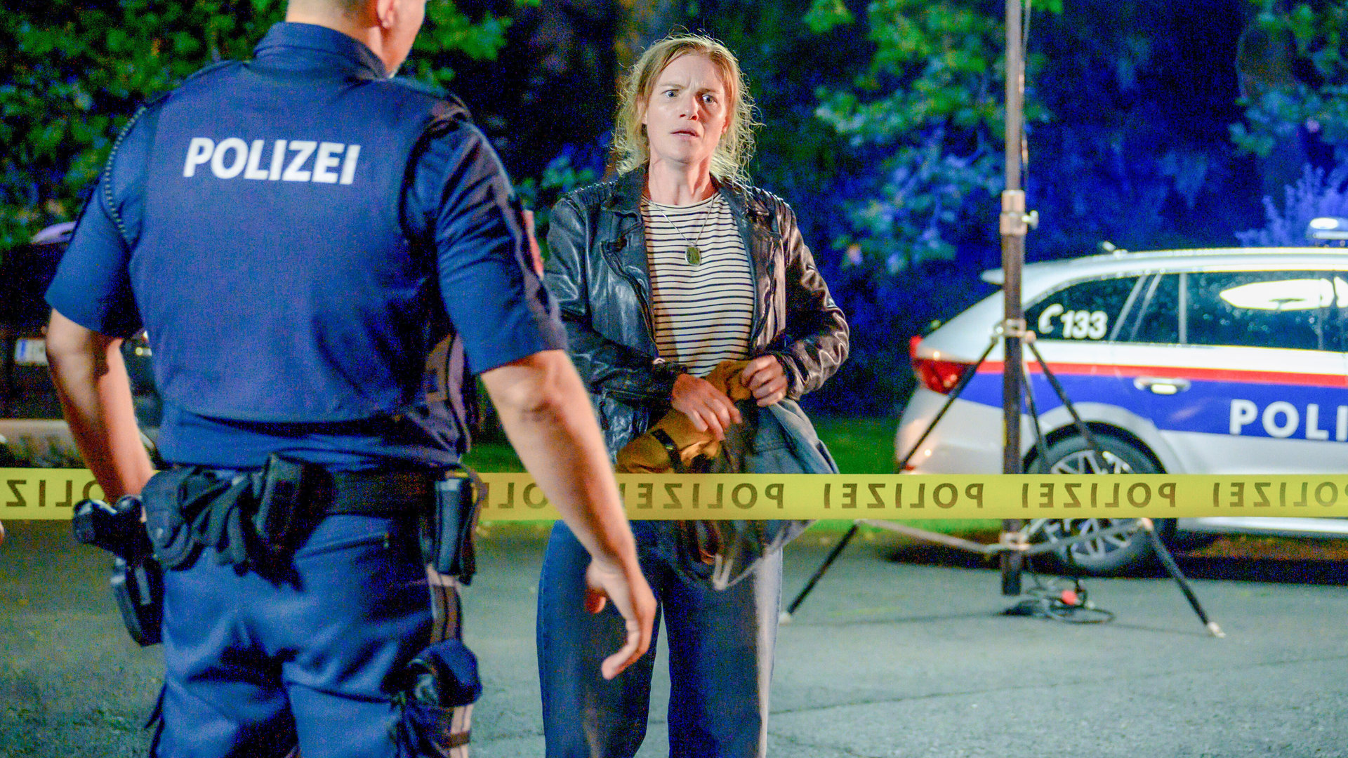 "SOKO Linz- Ist Luisa da?": Es ist Nacht aber Scheinwerfer und Blaulicht erhellen die Szene. Ein Polizist, den man von hinten sieht, bewacht das polizeiliche Absperrband, vor dem die verstörte, emotionale Esther Stadler (Michaela Schausberger) steht.