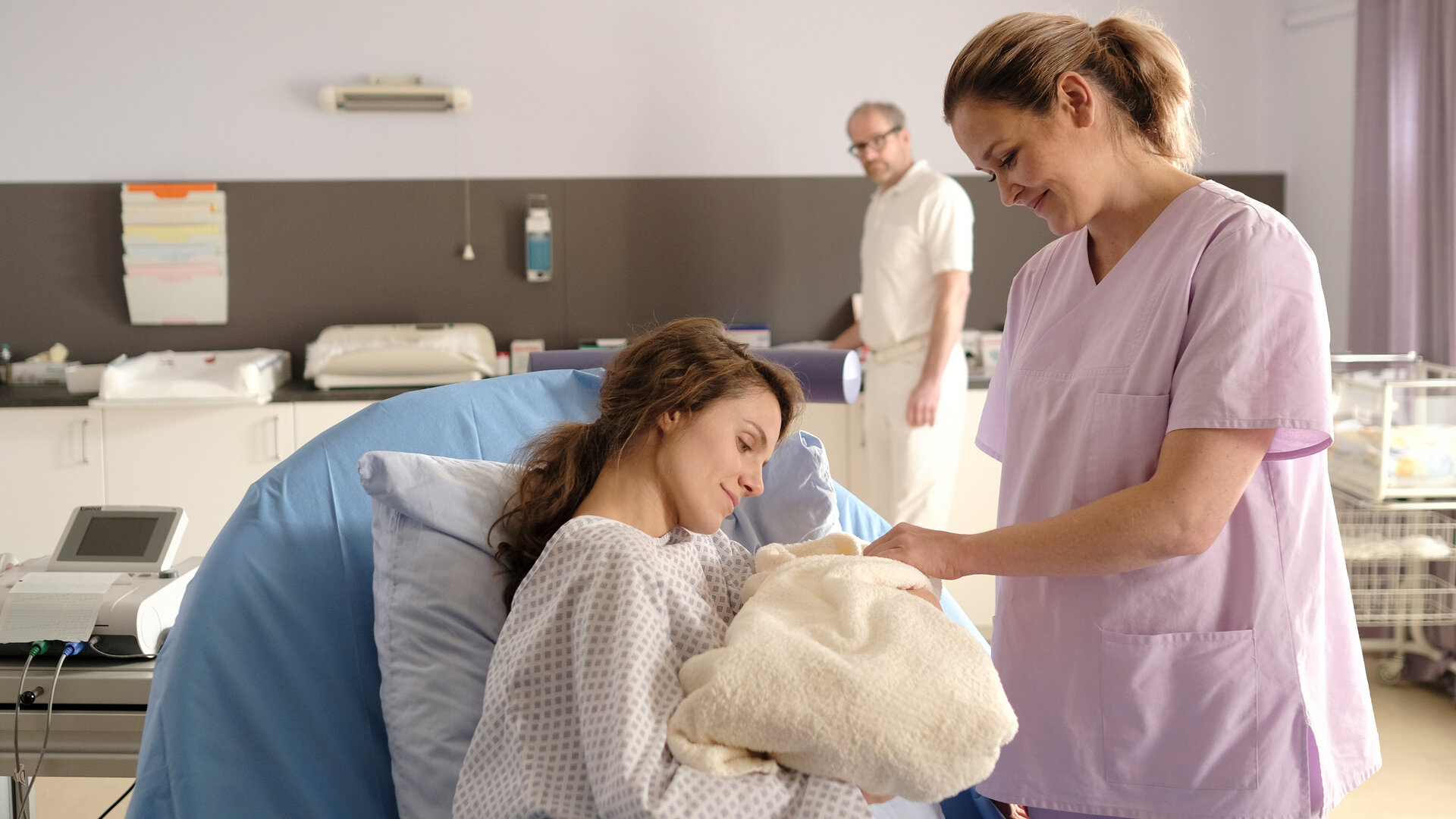 "Lena Lorenz - Hinter Gittern": Im Kreißsaal hält Sally Wilkens (Henriette Hölzel) ihr in Decken eingewickeltes Baby im Arm. Lena Lorenz (Judith Hoersch) steht neben ihr, während Dr. Keller (Thomas Limpinsel) die beiden von hinten beobachtet.