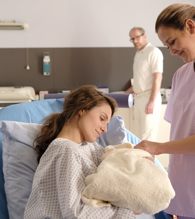 "Lena Lorenz - Hinter Gittern": Im Kreißsaal hält Sally Wilkens (Henriette Hölzel) ihr in Decken eingewickeltes Baby im Arm. Lena Lorenz (Judith Hoersch) steht neben ihr, während Dr. Keller (Thomas Limpinsel) die beiden von hinten beobachtet.