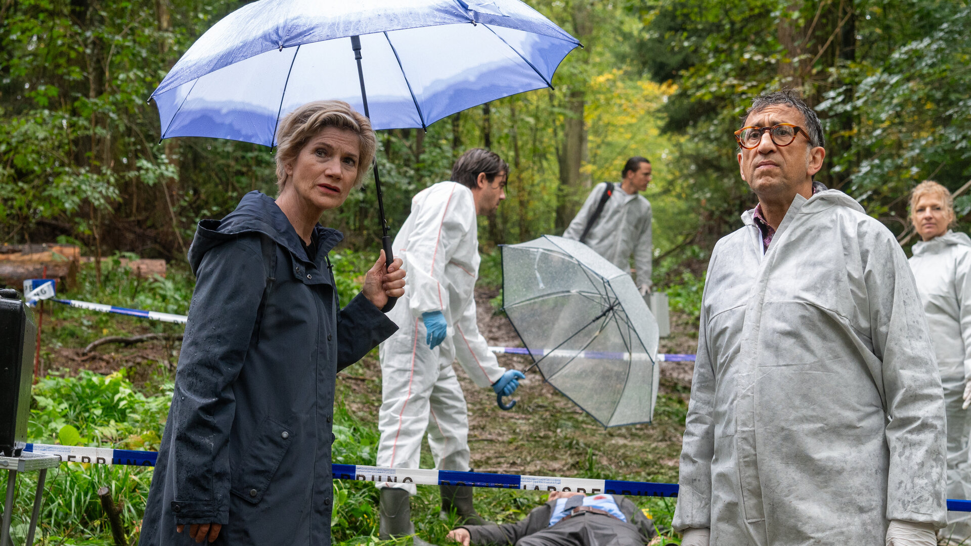 "SOKO Stuttgart - Waldesruh": Die erste Kriminalkommissarin Martina Seiffert (Astrid M. Fünderich) steht mit dem Leiter der KTU Jan Arnaud (Mike Zaka Sommerfeldt) am verregneten Tatort im Wald und sie schauen in die Ferne. Martina hält einen Regenschirm in der Hand. Hinter ihnen liegt die männliche Leiche (Komparse), bekleidet in einen grauen Businessanzug und Krawatte, auf dem Waldboden. Auf seiner Brust ist eine blutige Wunde. Der Leiter der Rechtsmedizin Dr. Benedikt Förster (Florian Wünsche) hält einen Regenschirm über den Leichnam. Weitere Mitarbeiter der Spurensicherung (Komparsen) arbeiten im Hintergrund.