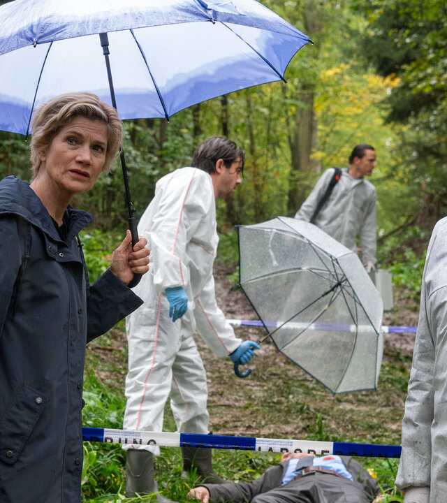 "SOKO Stuttgart - Waldesruh": Die erste Kriminalkommissarin Martina Seiffert (Astrid M. Fünderich) steht mit dem Leiter der KTU Jan Arnaud (Mike Zaka Sommerfeldt) am verregneten Tatort im Wald und sie schauen in die Ferne. Martina hält einen Regenschirm in der Hand. Hinter ihnen liegt die männliche Leiche (Komparse), bekleidet in einen grauen Businessanzug und Krawatte, auf dem Waldboden. Auf seiner Brust ist eine blutige Wunde. Der Leiter der Rechtsmedizin Dr. Benedikt Förster (Florian Wünsche) hält einen Regenschirm über den Leichnam. Weitere Mitarbeiter der Spurensicherung (Komparsen) arbeiten im Hintergrund.