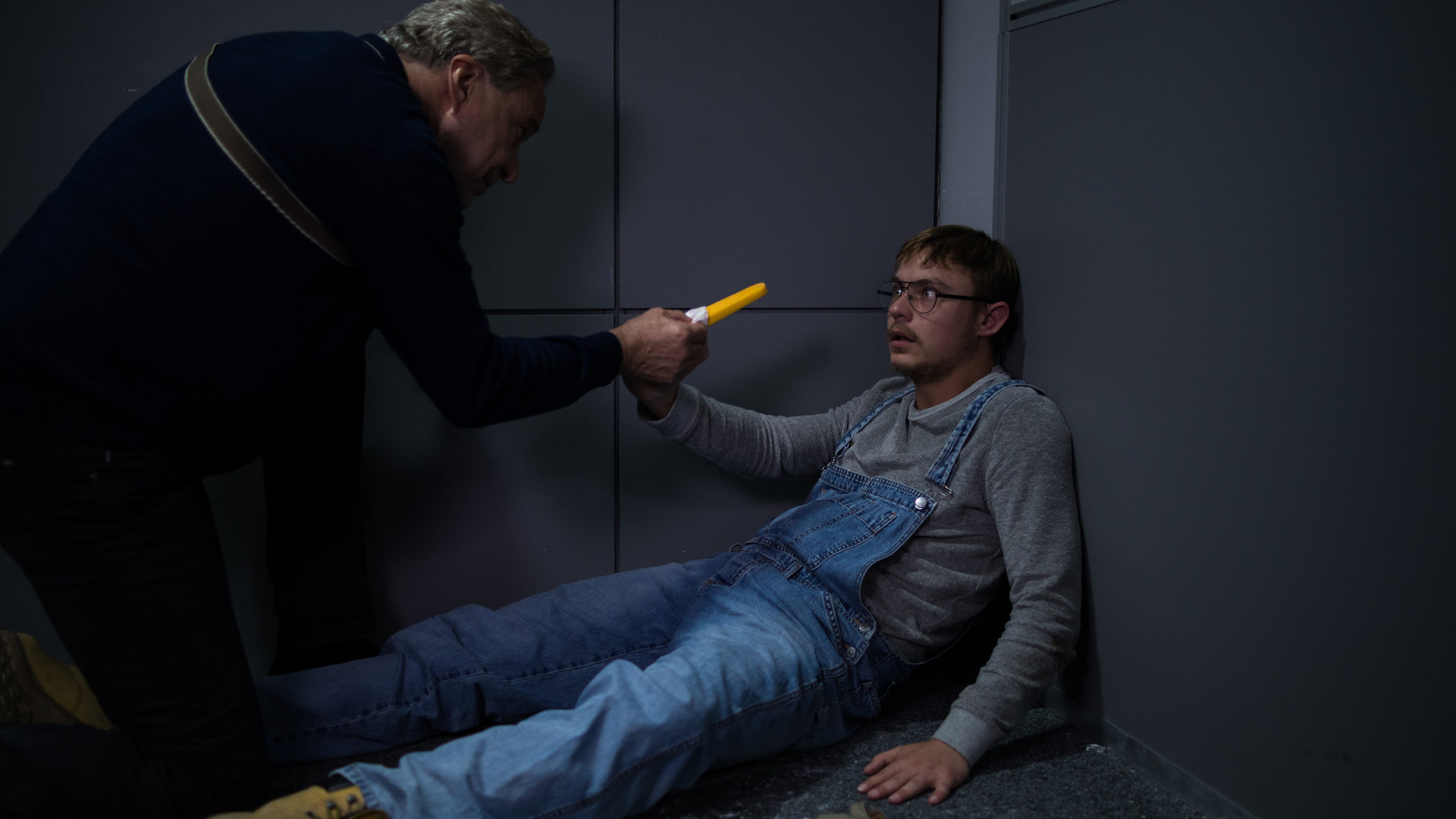 "SOKO München - Machtlos": Arthur Bauer (Gerd Silberbauer) kniet über Adam Klein (Dennis Mojen) im Verhörraum und streckt ihm ein Eis entgegen. Klein sitzt in der Ecke auf dem Boden und schaut Bauer mit großen Augen an.