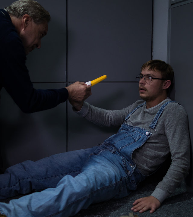 "SOKO München - Machtlos": Arthur Bauer (Gerd Silberbauer) kniet über Adam Klein (Dennis Mojen) im Verhörraum und streckt ihm ein Eis entgegen. Klein sitzt in der Ecke auf dem Boden und schaut Bauer mit großen Augen an.