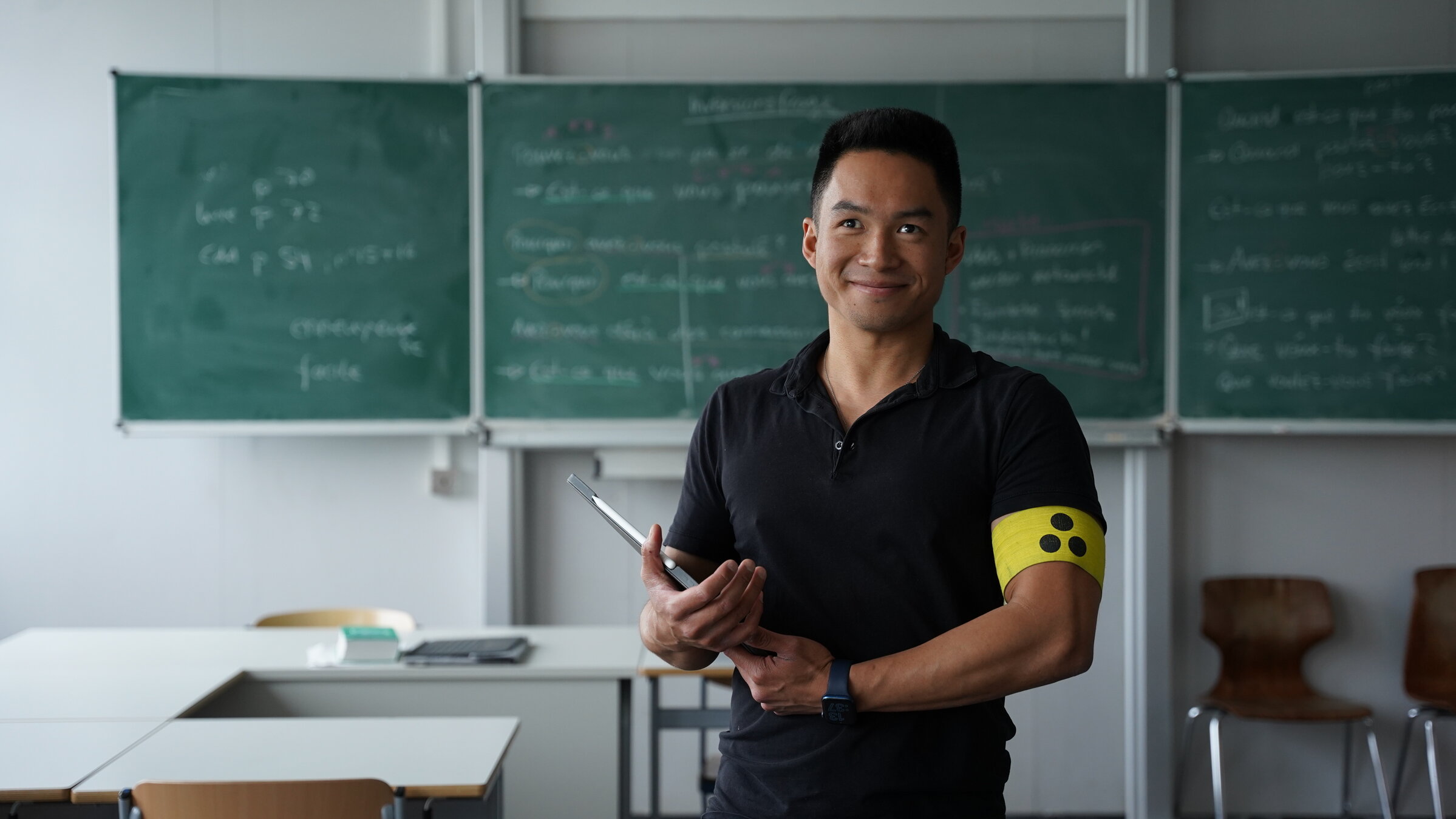 "Einfach Mensch! - Du bist mehr!": Wai Long Van steht mit einer gelben Armbinde mit drei schwarzen Punkten vor einer Schultafel. Er blickt lächelnd nach vorne.