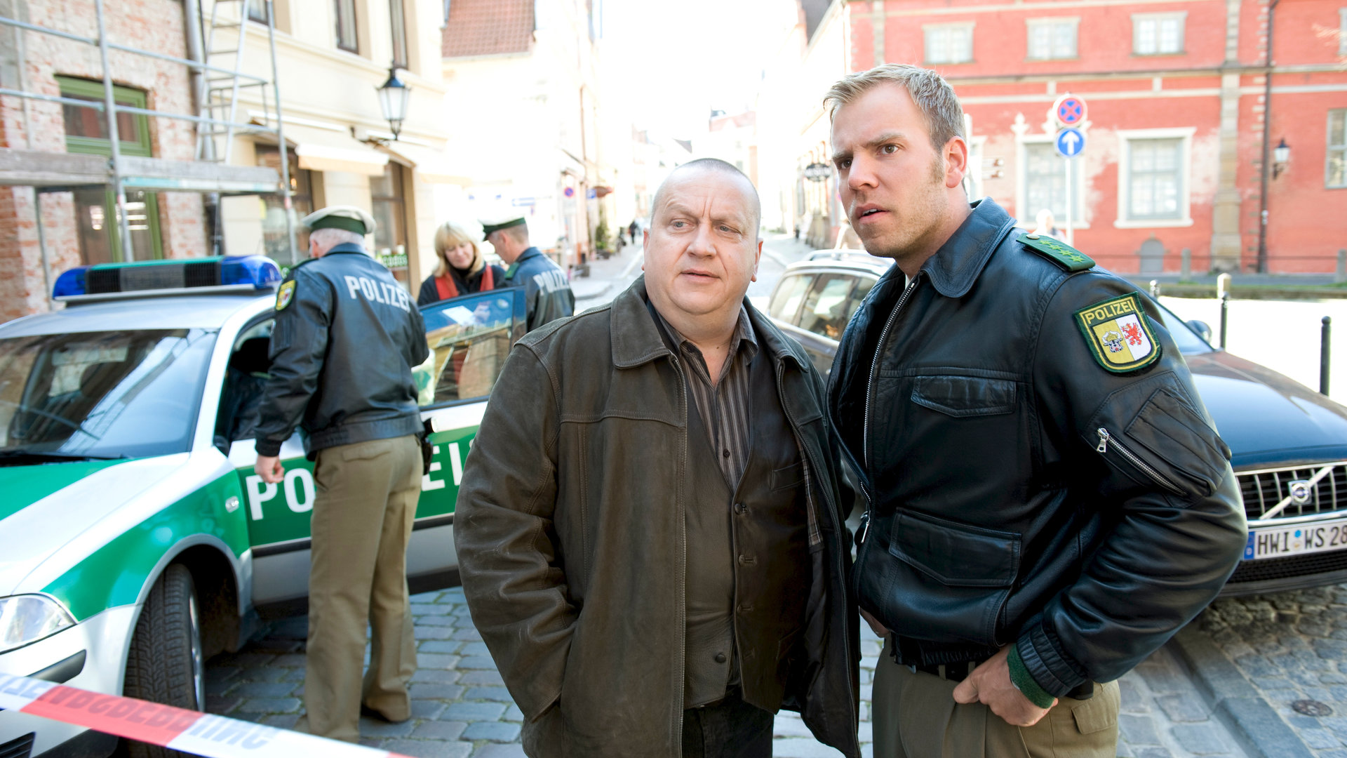"SOKO Wismar - Der Tod ist schneller": Jan Reuter (Udo Kroschwald) und Lars Pöhlmann (Dominic Boeer) stehen hinter der Polizeiabsperrung und beobachten etwas. Hinter ihnen sind Dienstfahrzeuge zu erkennen sowie die Innenstadt von Wismar.