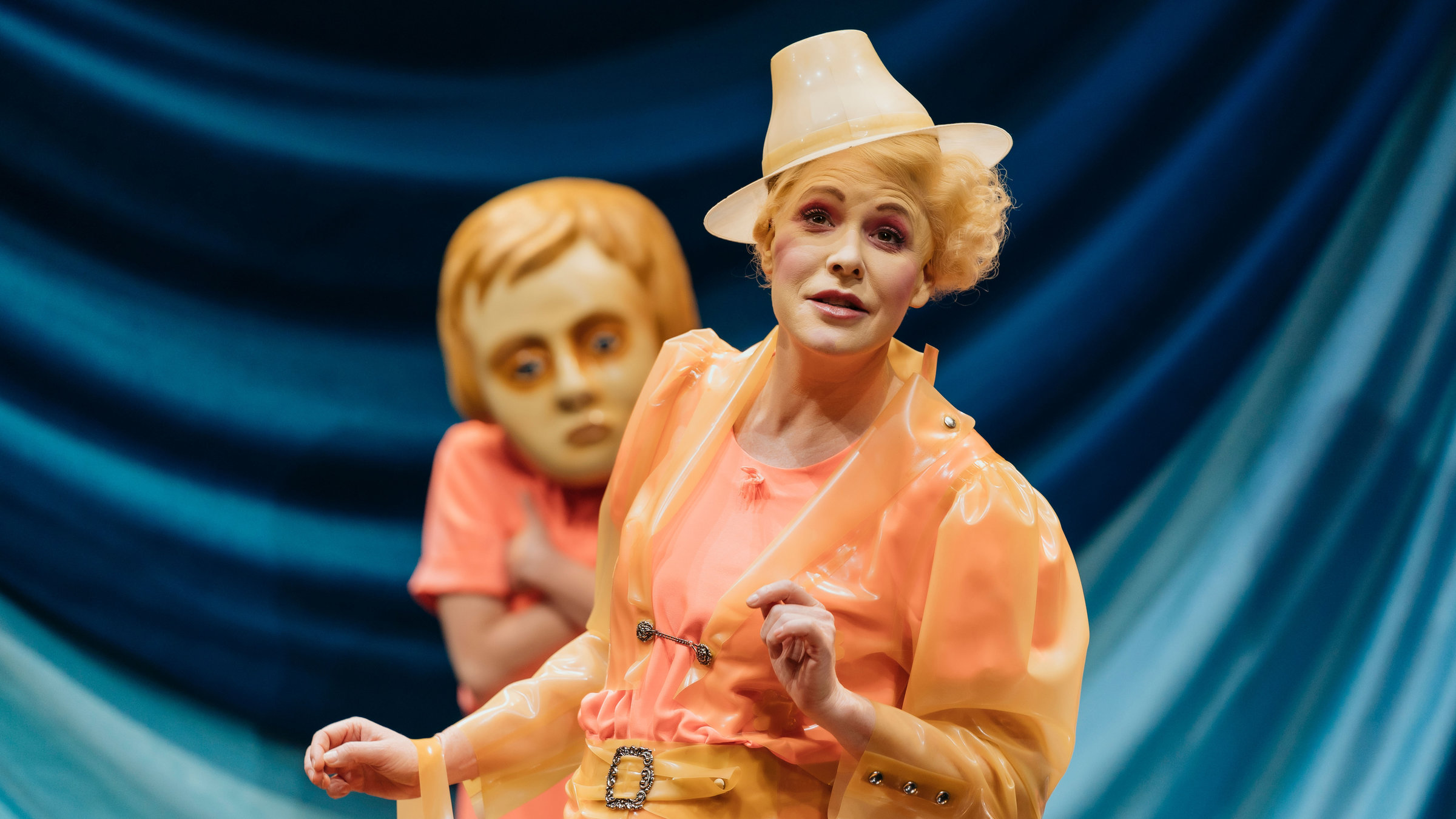 "Starke Stücke: Die Eingeborenen von Maria Blut": Eine schlanke Frau mit blondem welligem Haar blickt Richtung Publikum. Sie ist von der Hüfte aufwärts zu sehen und trägt ein Kostüm und einen kleinen Hut aus lachsfarbenem Latex im Stil von Trachtenmode. Hinter ihr steht eine Person, leicht unscharf, mit verschränkten Armen und einem übergroßen Puppenkopf. Die Bühnenrückwand ist mit einem leuchtend blauen Stoff verhängt.