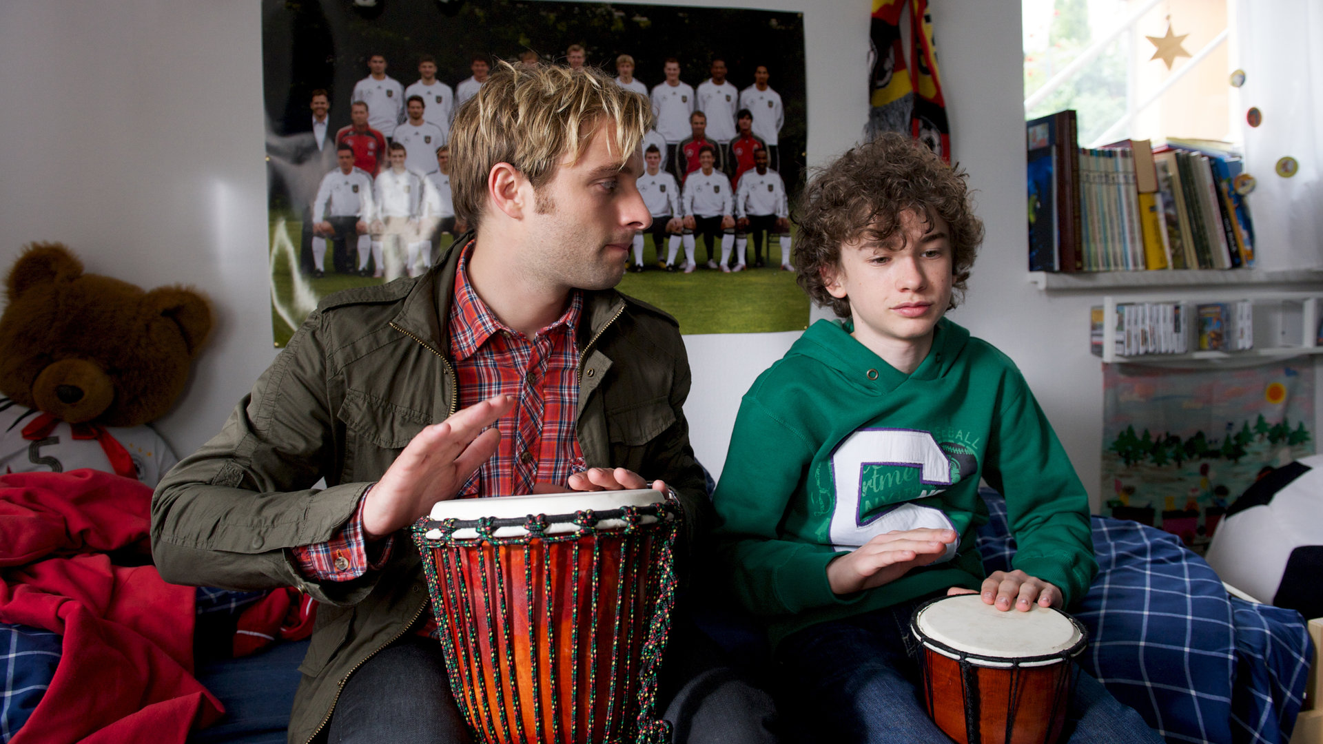 "SOKO Wismar - Wunderkind": Alex (Adrian Benjamin Moore) und Kommissar Nils Theede (Jonas Laux) sitzen in Alex´ Zimmer auf dem Bett nebeneinander mit Buschtrommeln auf dem Schoß. Im Hintergrund sind ein Fußballposter und Bücher.