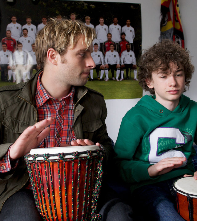 "SOKO Wismar - Wunderkind": Alex (Adrian Benjamin Moore) und Kommissar Nils Theede (Jonas Laux) sitzen in Alex´ Zimmer auf dem Bett nebeneinander mit Buschtrommeln auf dem Schoß. Im Hintergrund sind ein Fußballposter und Bücher.