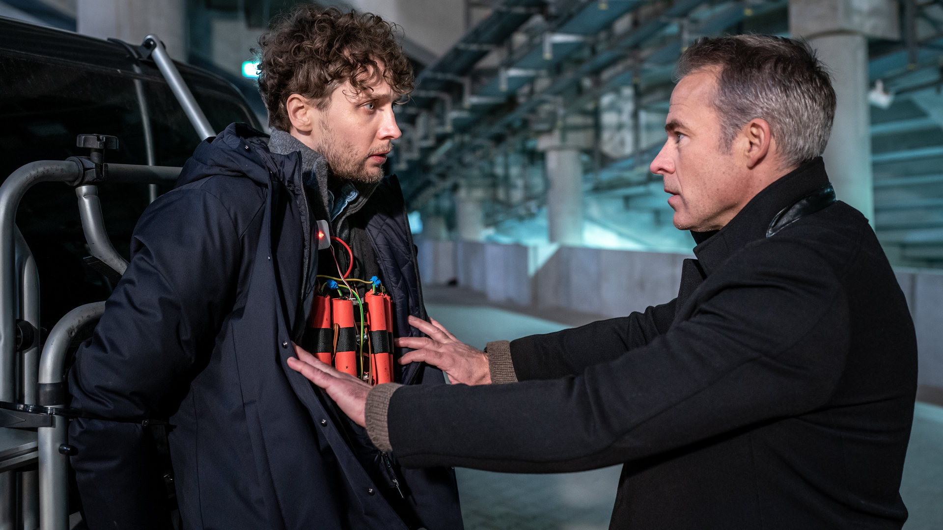 "SOKO Leipzig - Gegen die Zeit": Hanno Bieling (Julius Feldmeier) steht in einem Parkhaus, mit den Händen an den Kofferraum eines Vans gefesselt. Jan Maybach (Marco Girnth) steht ihm gegenüber und öffnet Bielings Jacke, unter der eine Sprengstoffweste zum Vorschein kommt. Die beiden Männer schauen sich entsetzt an.
