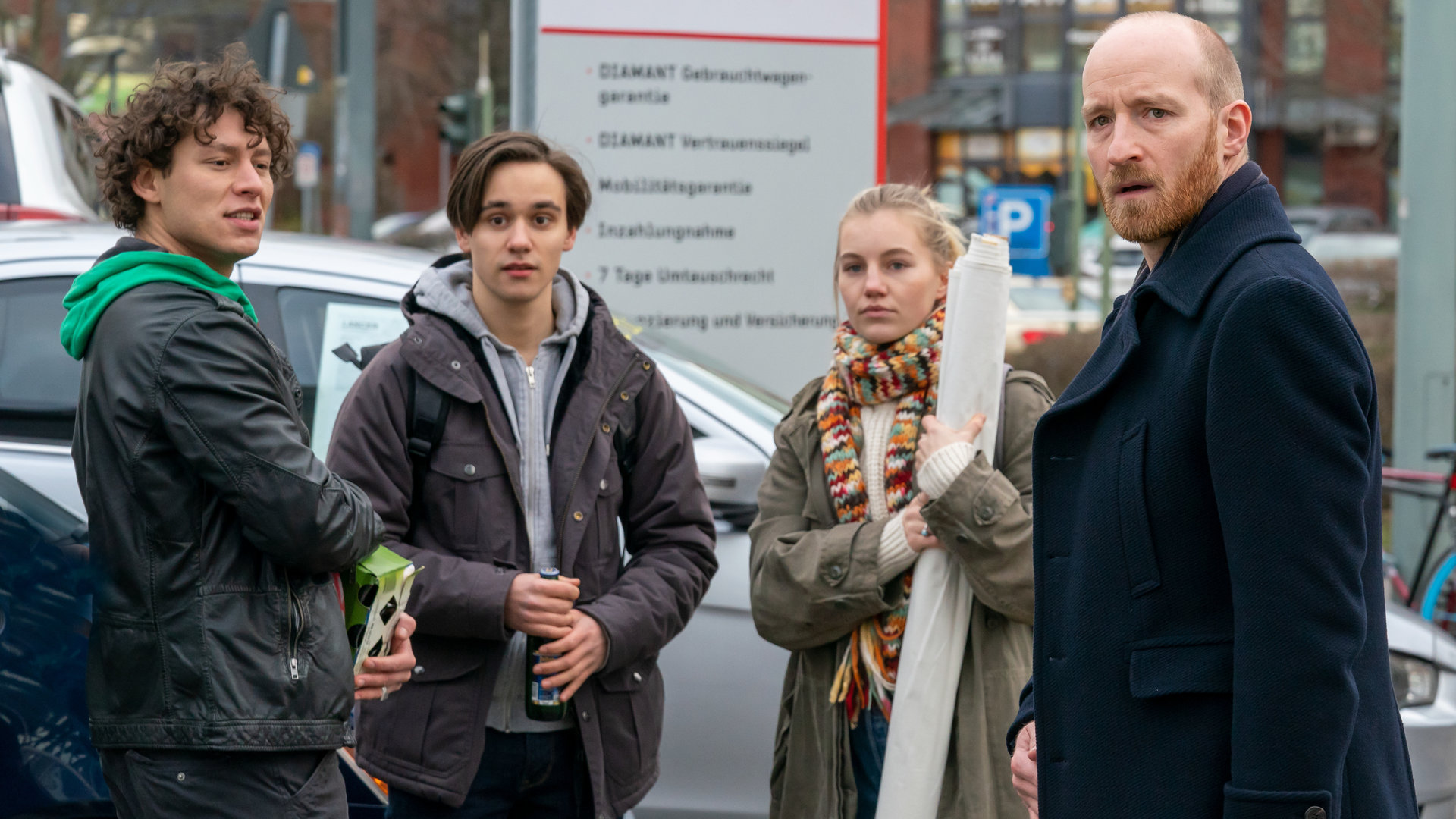 "Letzte Spur Berlin - Klimaangst": Finn Berger (Louie Betton), Daniel (Malik Blumenthal), Elli (Louise Sophie Arnold) und Jens Ehrenberg (Lukas Turtur) stehen vor einem Autohaus. Elli hält ein aufgerolltes Banner in der Hand, Finn eine Bierflasche.