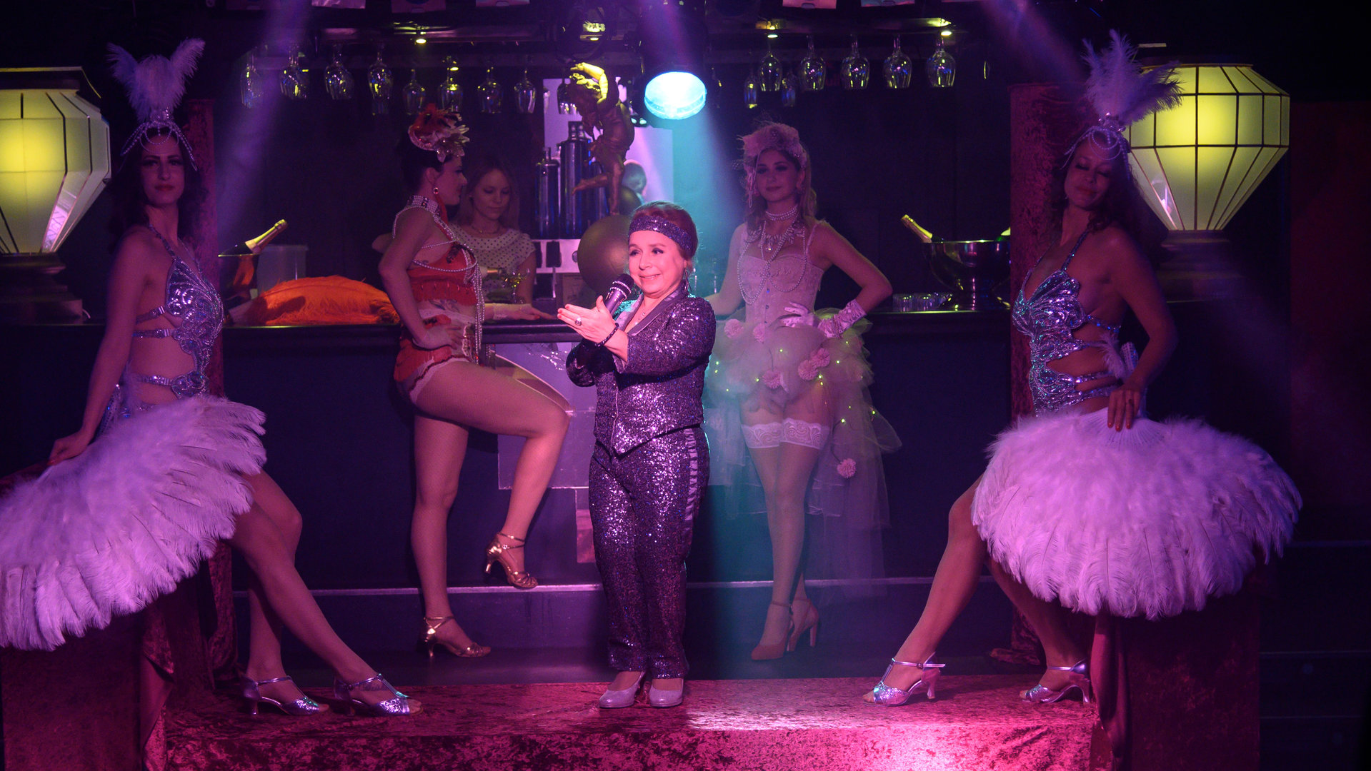 "SOKO Stuttgart – Burlesque": Dora Krumme (ChrisTine Urspruch) steht auf der Bühne des Burlesque-Clubs "Salon Rouge" auf der Bühne, die in schummriges Licht getaucht ist. Um sie herum sind vier Tänzerinnen (Komparsinnen) in erotischen Burlesque-Outfits. Sie haben etwa Federnschmuck auf dem Kopf und um die Hüfte, tragen Ketten sowie glänzende Pumps. Zwei von ihnen stehen an einer Bar, die man im Hintergrund sieht, die anderen beiden sitzen links und rechts auf der Bühne auf einer Art Geländer. Krumme ist in einen glitzernden Anzug gekleidet, hält ein Mikrofon in der Hand und macht eine präsentierende Geste in Richtung Publikum.