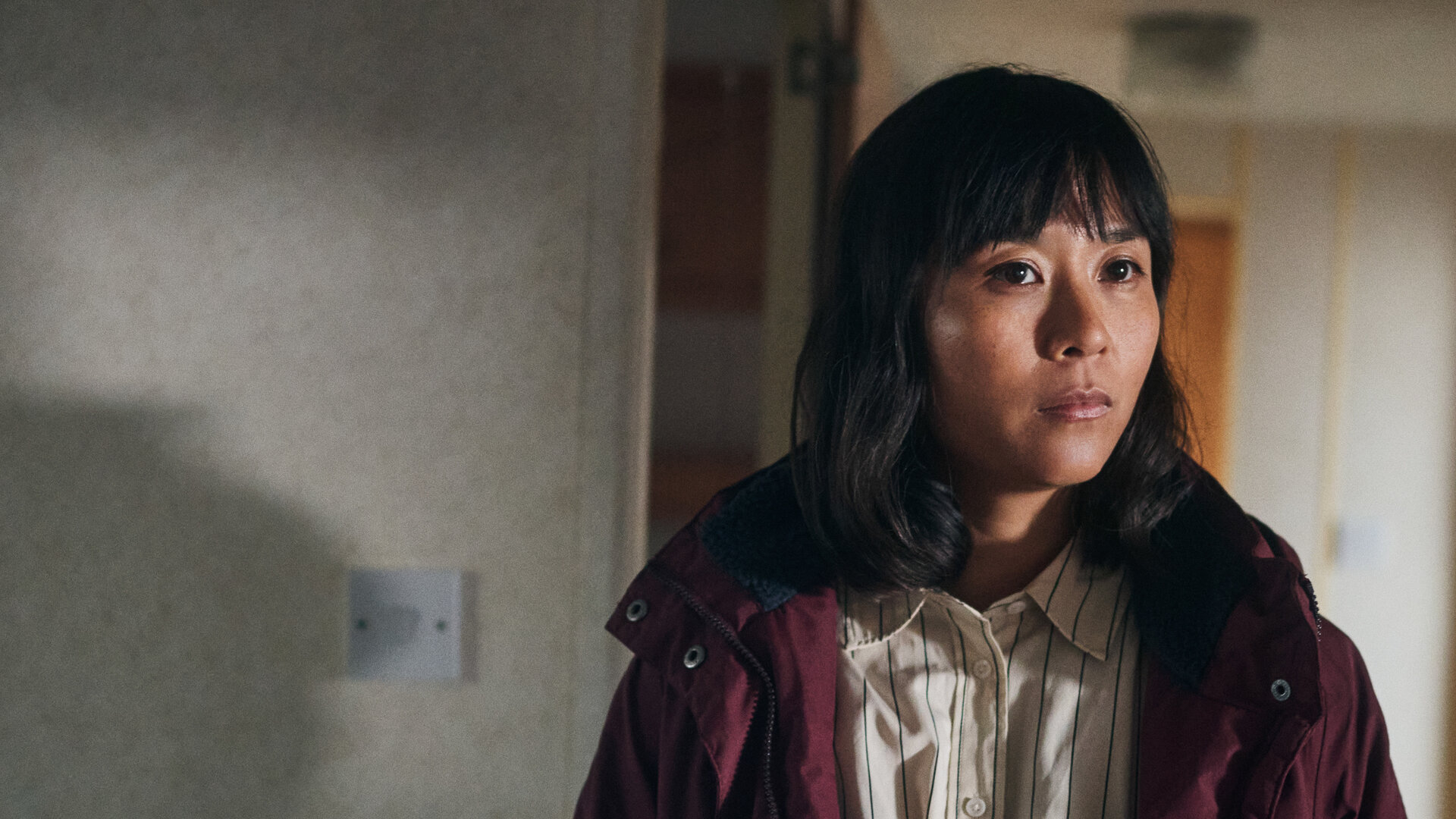 "Under Salt Marsh - Tod in den Salzwiesen - Die schwarze Sau": Jess (Naomi Yang) steht in einem Raum. Sie trÃ¤gt einen Mantel und blickt nach vorne.