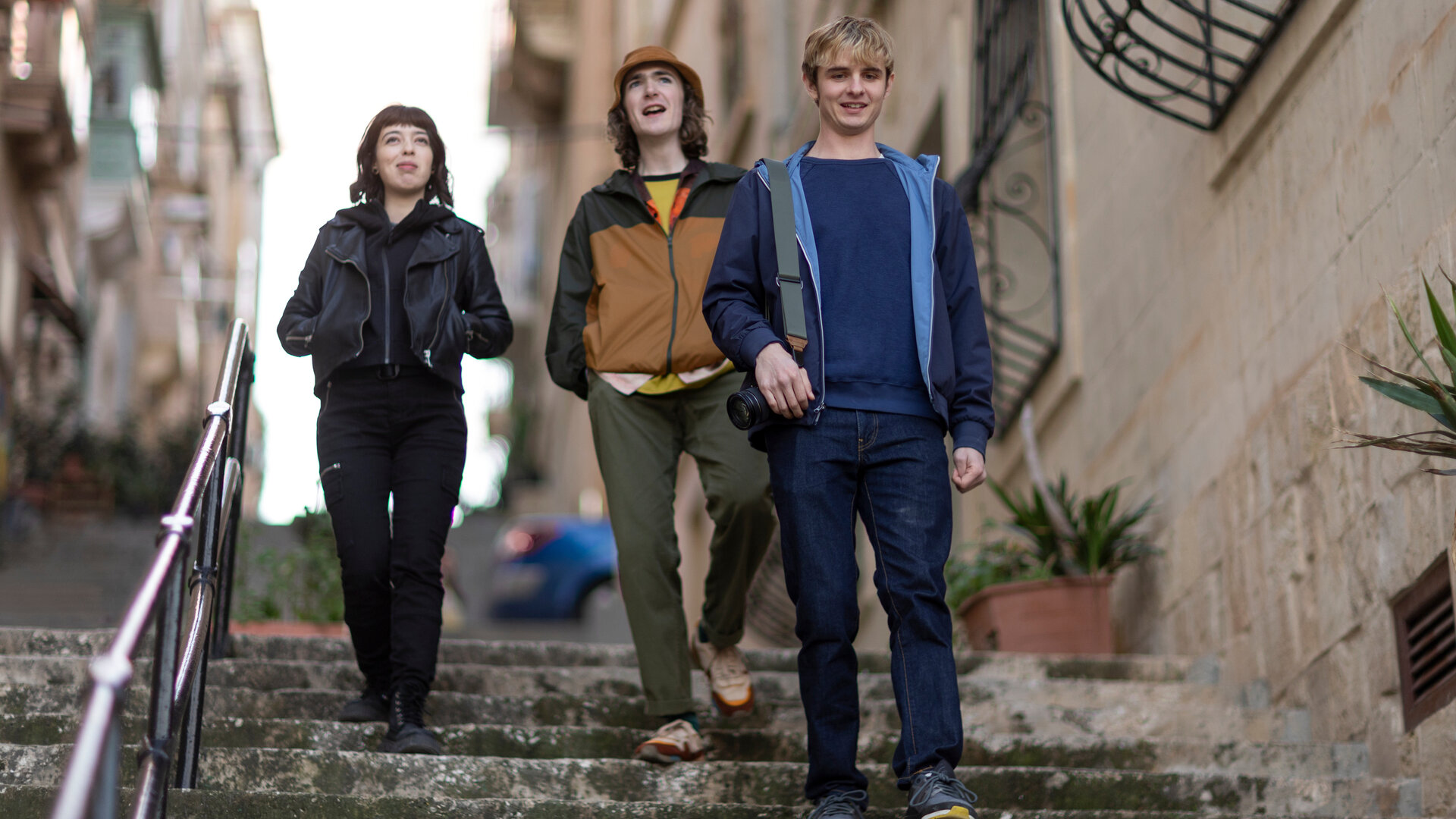 "Alex Rider - Witwe": Alex Rider (Otto Farrant) geht mit einem Fotoapparat in der Hand eine steinerne Treppe hinab, gefolgt von seinen Freunden Tom (Brenock O'Connor) und Kyra (Marli Siu). Sie befinden sich in einer engen, von historischen Gebäuden gesäumten Gasse mit Pflanzen und Balkongittern.