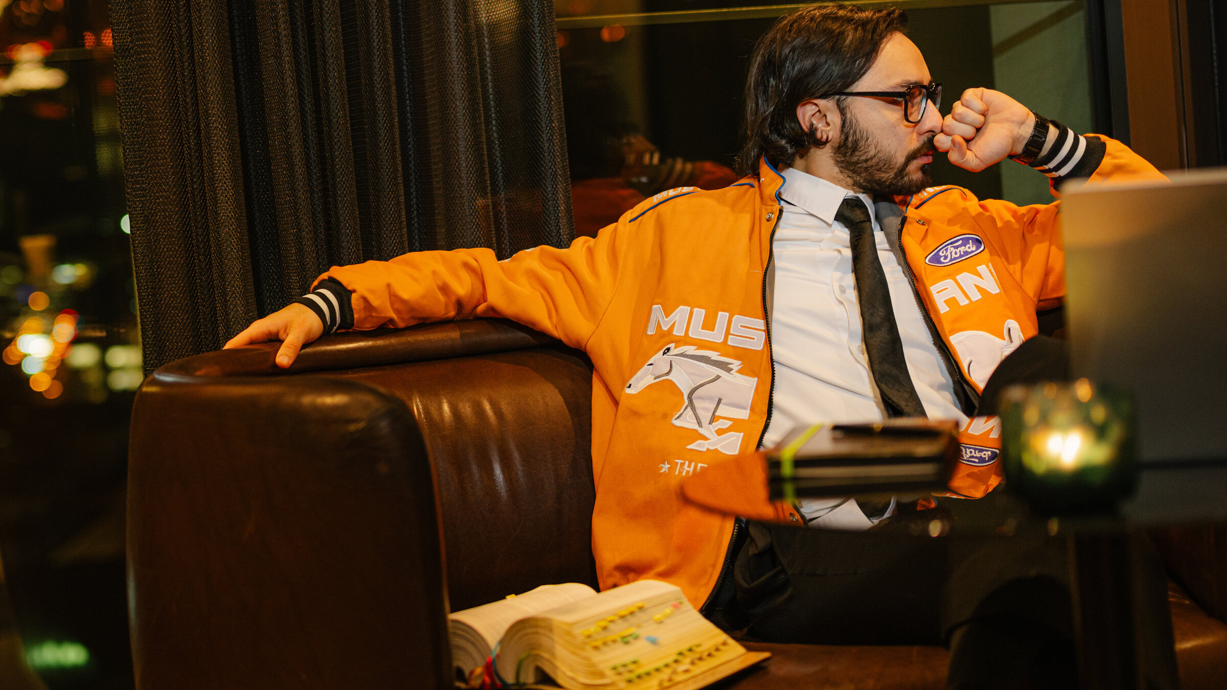 "37° Leben: PHASE: Alles Krise": Furkan, ein junger Mann, sitzt auf einer braunen Couch und schaut zur Seite. Neben ihm liegt ein dickes, aufgeschlagenes Buch mit farbigen Klebezetteln.