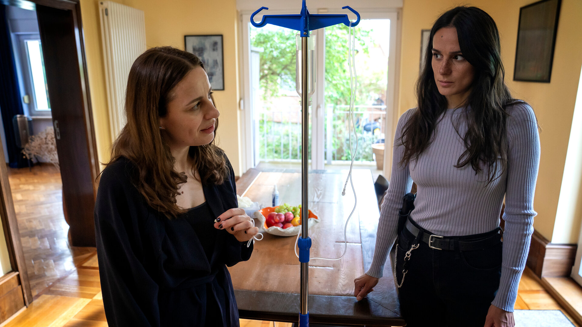 "Jenseits der Spree - Lieferkosten inklusive": Christine Margau (Gina Henkel) steht  Mavi Neumann (Aybi Era) in einem Zimmer gegenüber. Zwischen den beiden steht ein Tropf, an den Christine Margau angeschlossen ist.