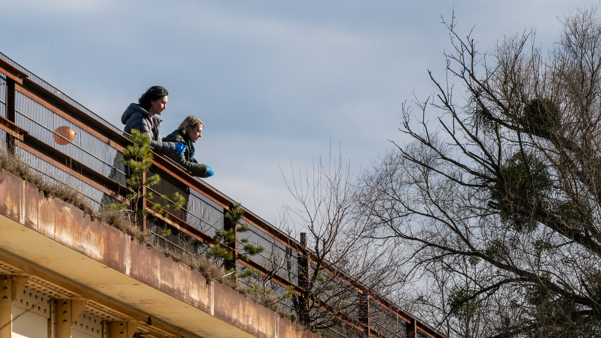 "SOKO Potsdam - Schwerelos": Auf einer alten, rostigen Brücke stehen die Kommissarinnen Pauline Hobrecht (Agnes Decker) und Tamara Meurer (Anja Pahl). Sie lehnen am Geländer und blicken hinunter. Beide wirken nachdenklich. Über ihnen ist der Himmel graublau.