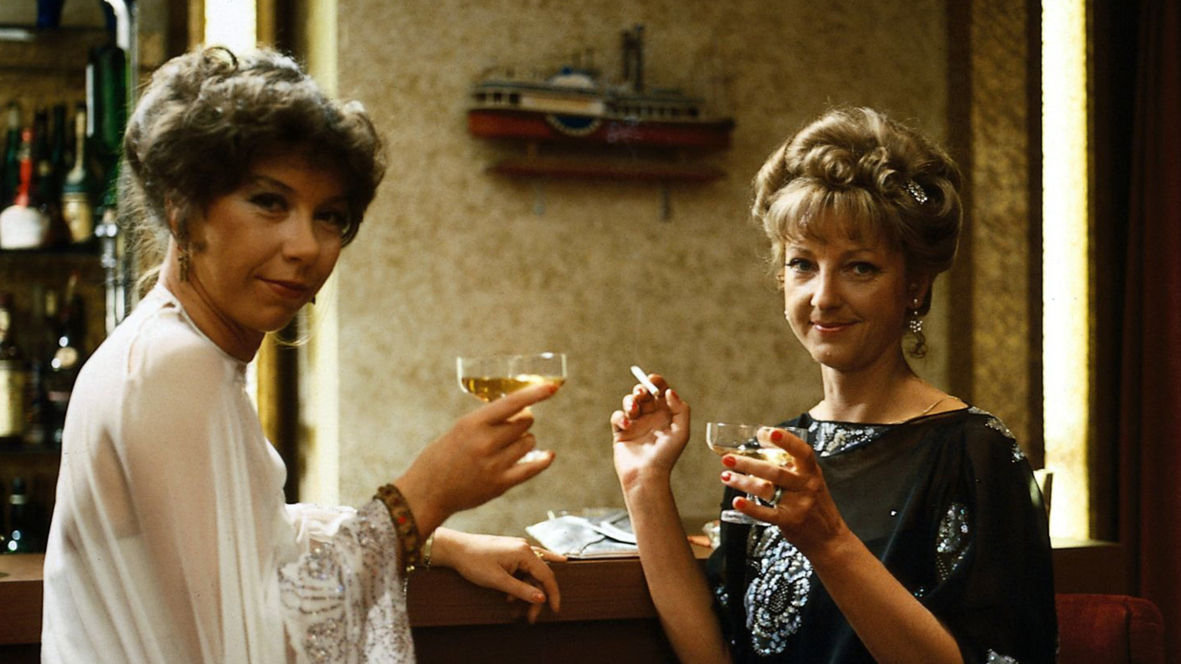 "Das Traumschiff": Die beiden edlen Damen Frau von Schall (Evelyn Hamann) und Frau Karsky (Corinna Genest) sitzen im Zimmer vor einem Spiegel, lächeln in die Kamera und trinken Champagner. Frau Karsky hält eine Zigaretten in der rechten Hand.