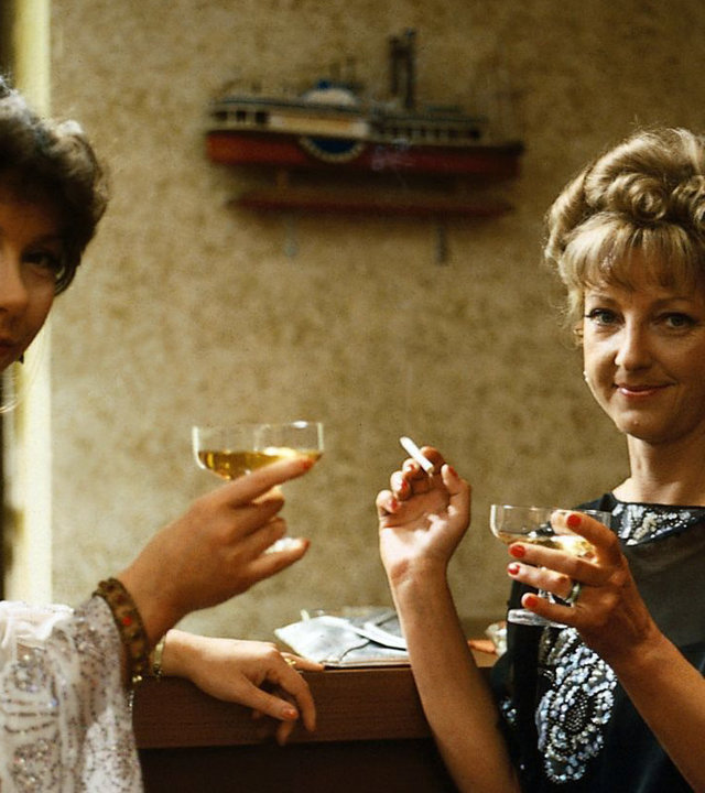 "Das Traumschiff": Die beiden edlen Damen Frau von Schall (Evelyn Hamann) und Frau Karsky (Corinna Genest) sitzen im Zimmer vor einem Spiegel, lächeln in die Kamera und trinken Champagner. Frau Karsky hält eine Zigaretten in der rechten Hand.