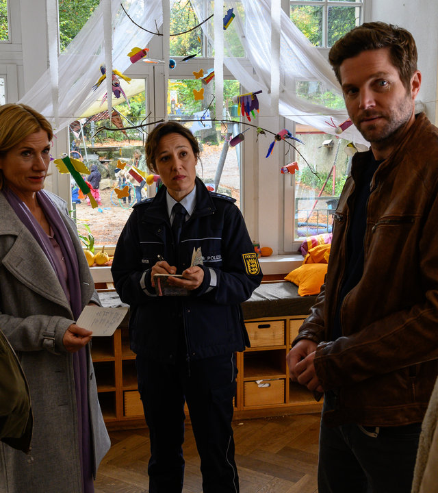 "SOKO Stuttgart – Kindersegen": Ines Richter (Davina Donaldson), Martina Seiffert (Astrid M. Fünderich), Sibylle Beyer (Bärbel Stolz) und Joachim Stoll (Peter Ketnath) stehen in einem Raum der Kindertagesstätte zusammen und schauen alle interessiert auf Johann Kammerer (Carsten Strauch). Martina hält einen Zettel in den Händen. Im Hintergrund Regale mit Spielsachen und ein großes Fenster, vor dem ein buntes Mobile hängt.
