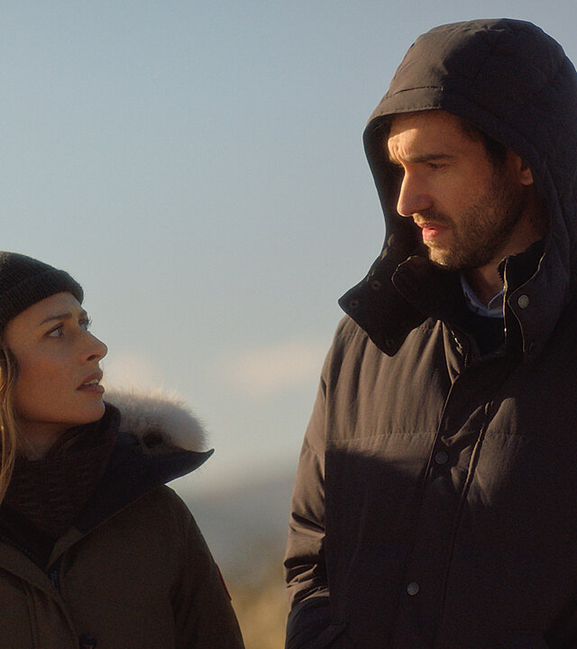 "Queenstown Murders – Zum Sterben schön - Aus heiterem Himmel": Detective Sergeant Anais Mallory (Chelsie Preston Crayford) und Detective Simon Delaney (Matt Whelan) stehen nebeneinander im Freien. Sie schauen sich gegenseitig an. Beide tragen dicke Jacken, Anais dazu eine Wollmütze. Simon hat seine Kapuze über den Kopf gezogen.