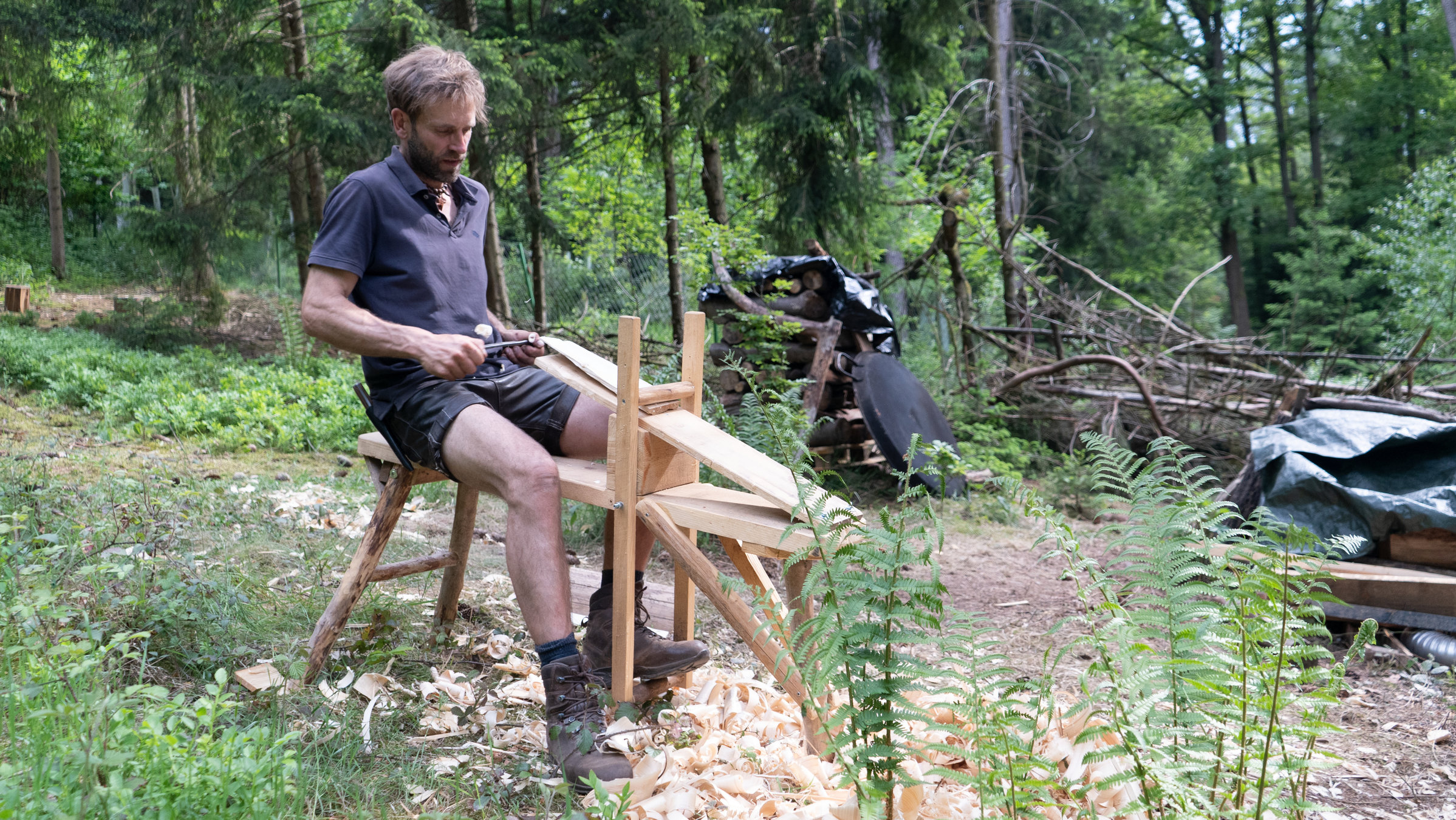 "37° Zuhause im Wald - Leben abseits der Zivilisation": Marc sitzt an einer selbstgebauten Werkbank und produziert Holzschindeln für das Dach seines Hauses. Im Hintergrund Wald.