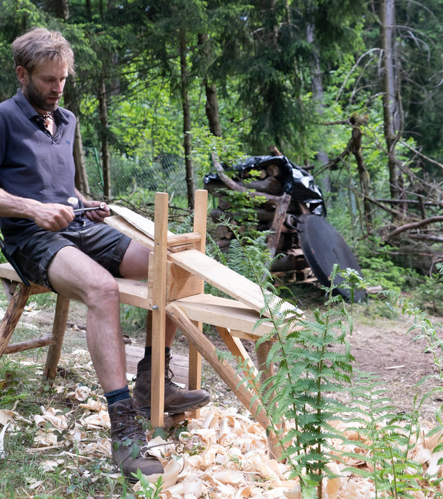"37° Zuhause im Wald - Leben abseits der Zivilisation": Marc sitzt an einer selbstgebauten Werkbank und produziert Holzschindeln für das Dach seines Hauses. Im Hintergrund Wald.