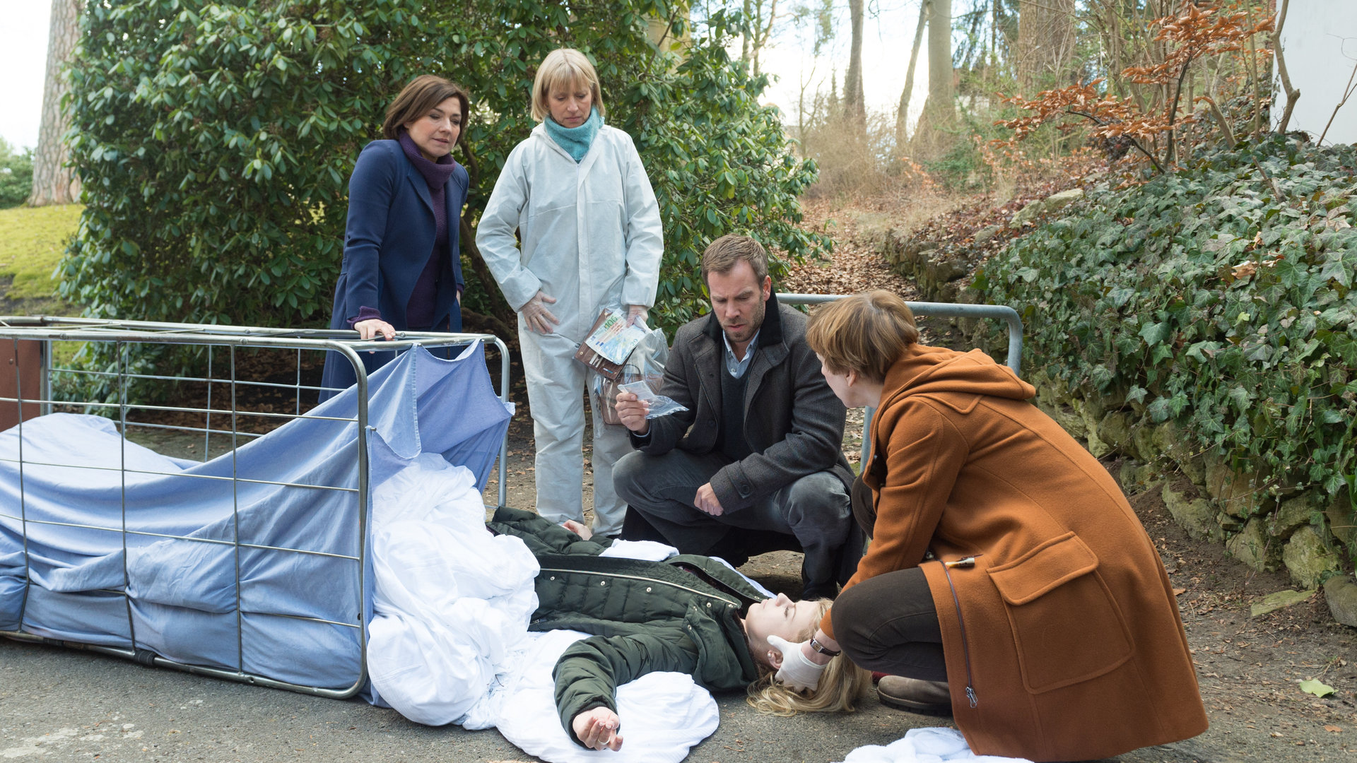 "SOKO Wismar – Die Freuden des Alters": Katrin Börensen (Claudia Schmutzler), Roswitha Prinzler (Silke Matthias), Lars Pöhlmann (Dominic Boeer) und Dr. Helene Sturbeck (Katharina Blaschke) stehen um einen umgekippten Wäschewagen herum, in dem zwischen Bettlaken eine weibliche Leiche liegt. Sturbeck untersucht den Kopf der Toten (Jenny Edner).