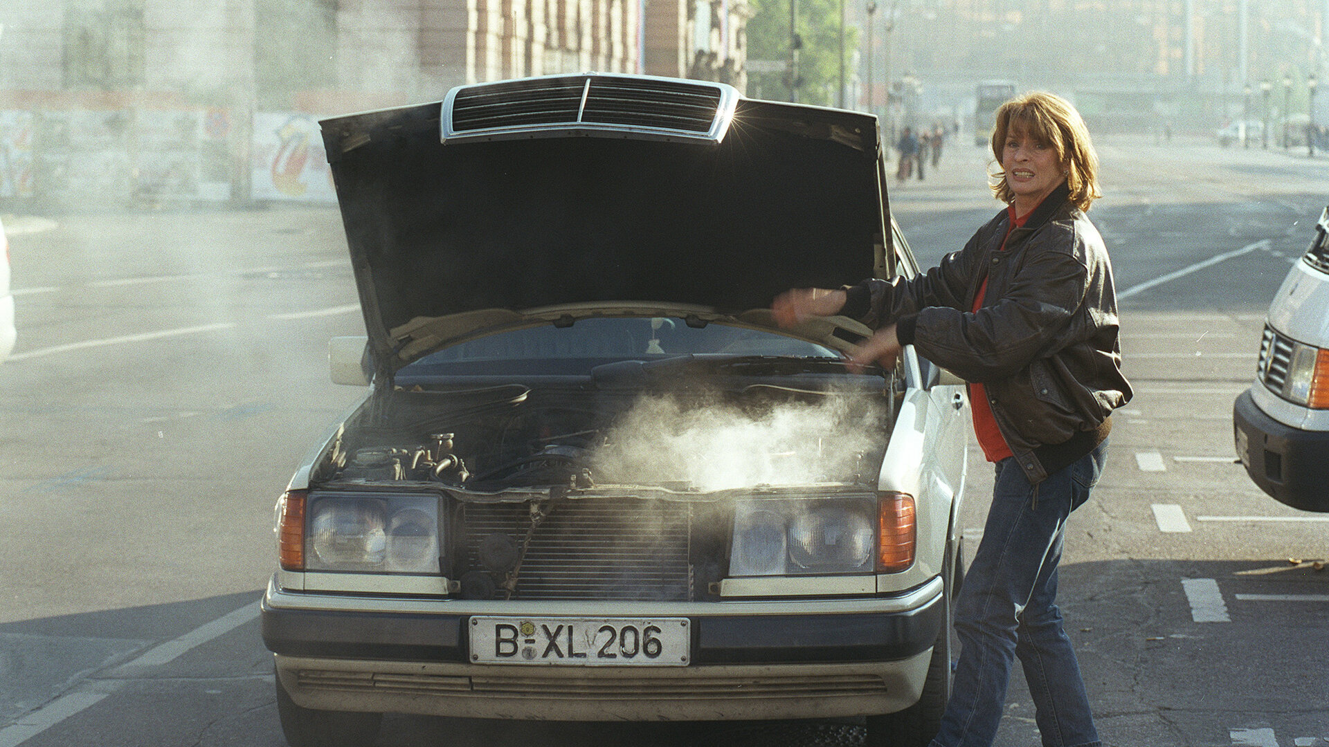 "Die schnelle Gerdi und die Hauptstadt – Die Tierschützerin": Gerdi (Senta Berger) steht an der Straße neben einem Taxi mit geöffneter Motorhaube. Aus dem Motorraum steigt Rauch auf, den sie mit den Händen zu vertreiben versucht.