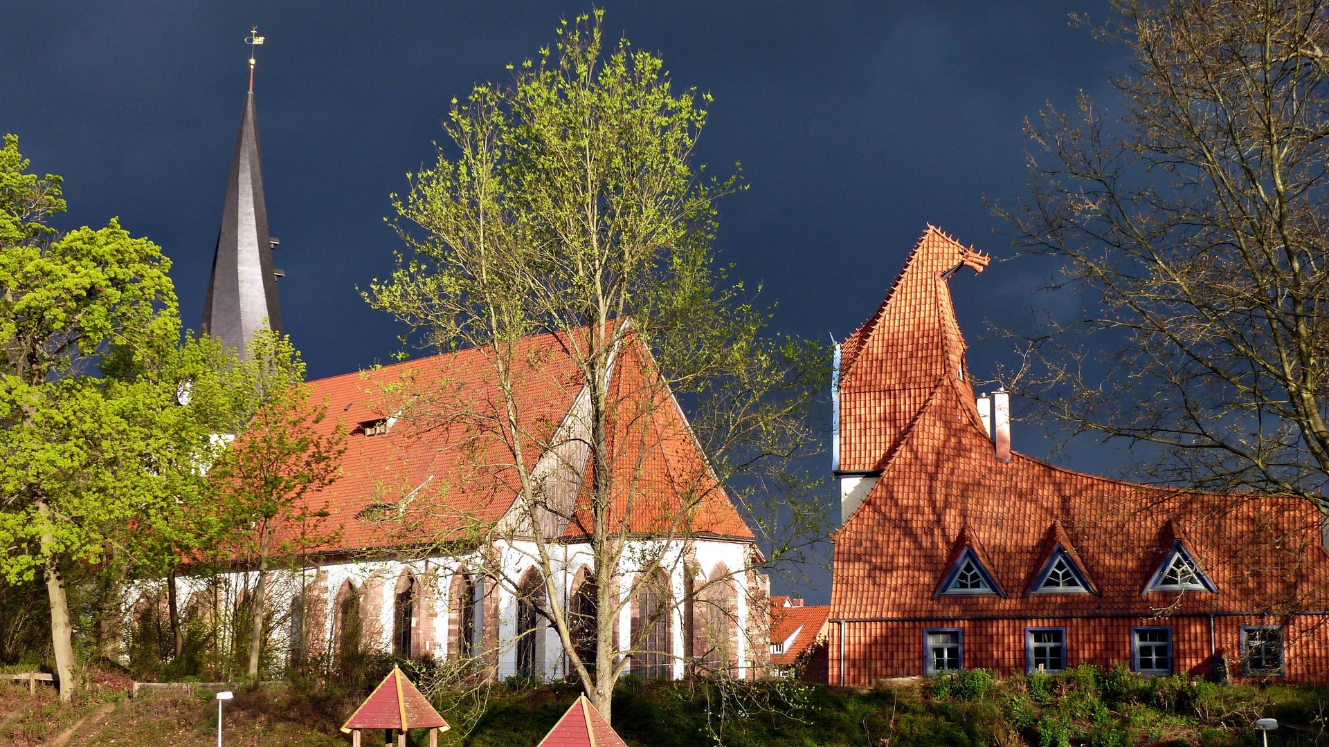 "Evangelischer Gottesdienst - Gute Nachricht für Alltagshelden": Außenansicht der filigranen Kirche St. Sixti in Northeim. Das Gebäude ist weiß mit vielen hohen Fenstern, das Dach ist mit hellroten Ziegeln gedeckt. Neben der Kirche befindet sich das Theater der Nacht, das architektonisch der Kirche angelglichen ist. Umgeben sind beide Gebäude von Bäumen mit hellgrünen Blättern.