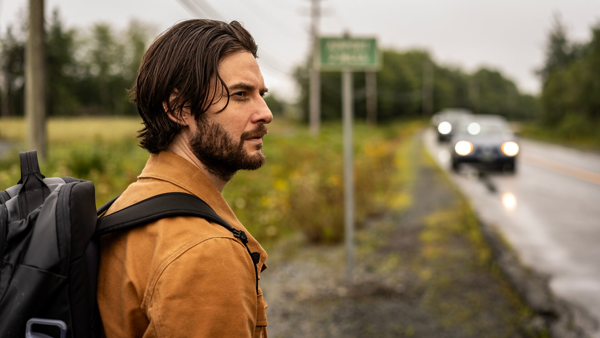 "Das Institut - Die Entführung": Mit einem Rucksack auf dem Rücken steht Tim Jamieson (Ben Barnes) an einer Landstraße. Im Hintergrund fahren in der Unschärfe Autos auf der Straße, daneben steht ein Straßenschild.