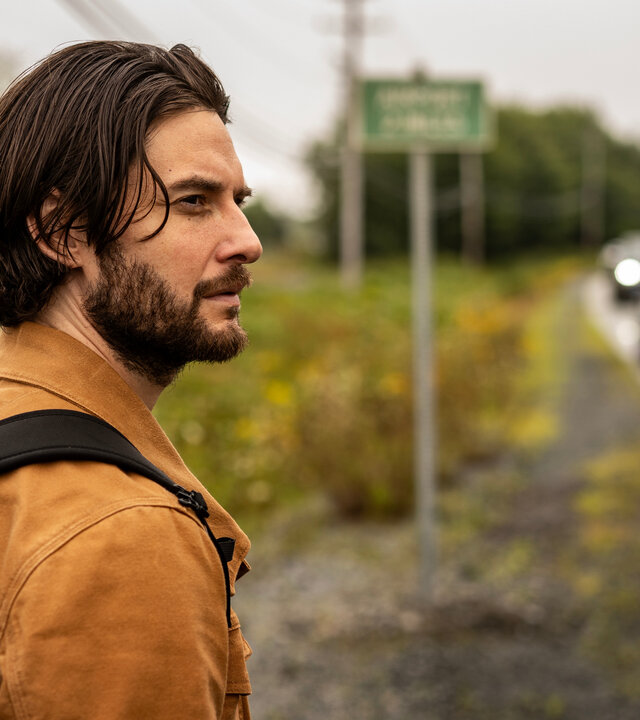 "Das Institut - Die Entführung": Mit einem Rucksack auf dem Rücken steht Tim Jamieson (Ben Barnes) an einer Landstraße. Im Hintergrund fahren in der Unschärfe Autos auf der Straße, daneben steht ein Straßenschild.