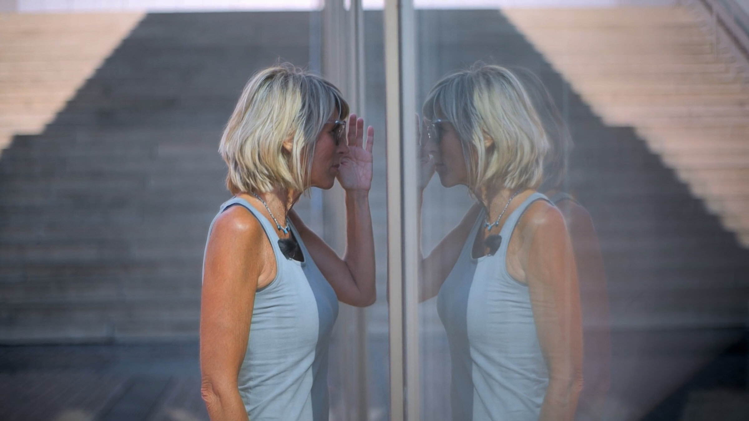 "37°: Die Entdeckung der Hoffnung - An Lebenskrisen wachsen": Steffi, eine sportlich schlanke Frau mit schulterlangen blonden Haaren steht an einer Glasscheibe und schaut hinein. Sie trägt eine Sonnenbrille und ein blaues Tank-Top. Neben ihr ist ihr Spiegelbild zu sehen.