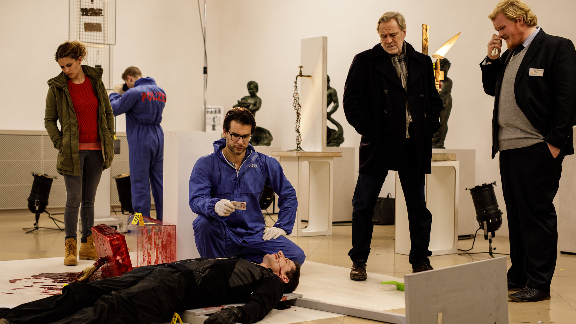 "SOKO München - Nachtwache": Am Tatort in einem Museum liegt eine Leiche am Boden , Dr. Weissenböck (Florian Odendahl) sitzt in der Hocke daneben und schaut sich den Ausweis der Leiche an. Rechts von der Leiche steht Antonia Bischoff (Amanda da Gloria), rechts steht Arthur Bauer (Gerd Silberbauer) neben ihm steht Jean-Claude (Sebastian Griegel) ein Angestellter des Museum.