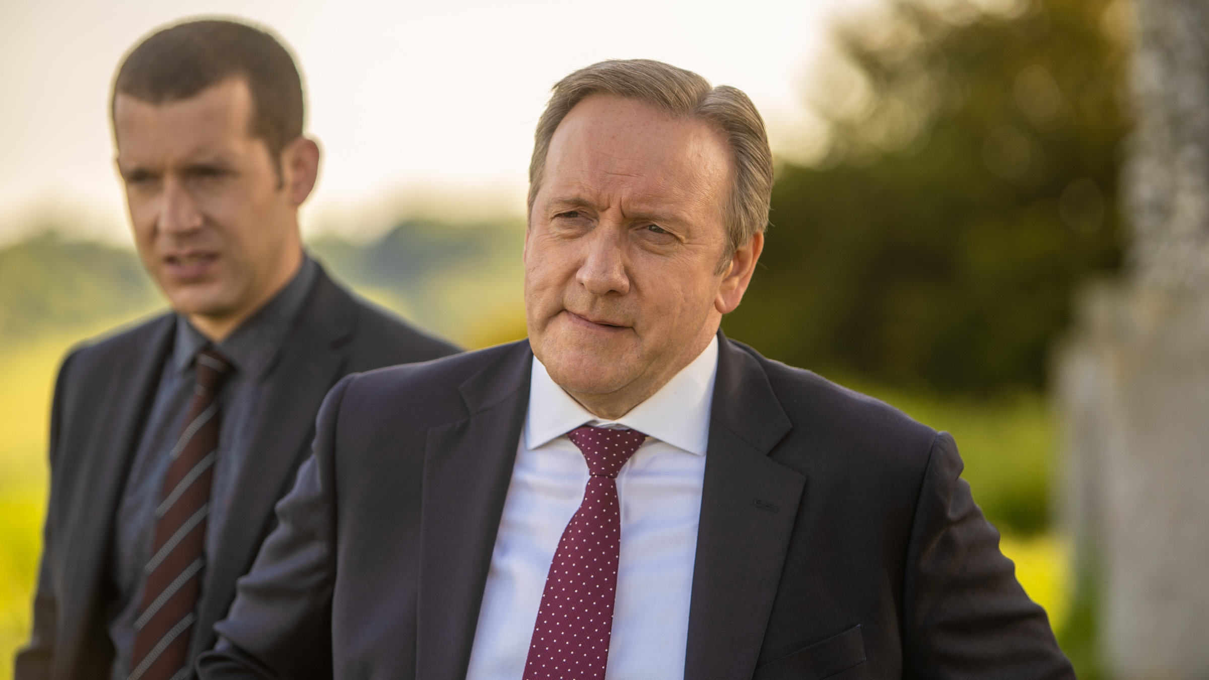 "Inspector Barnaby: Mord ist keine Hexerei": DS Jamie Winter (Nick Hendrix) und DCI John Barnaby (Neil Dudgeon) stehen in Anzügen gekleidet nebeneinander im Freien und schauen ernst nach links aus dem Bild.