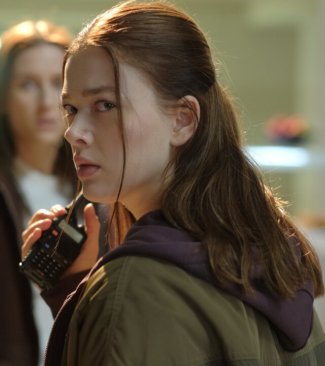 "Fallen - Ausweglos": Luce (Jessica Alexander) steht in einer Kantine und hält ein Funkgerät in der Hand, während sie mit angespanntem Blick zur Seite schaut. Hinter ihr sind Arianne (Josefine Koenig) und Molly (Maura Bird) leicht unscharf zu erkennen.