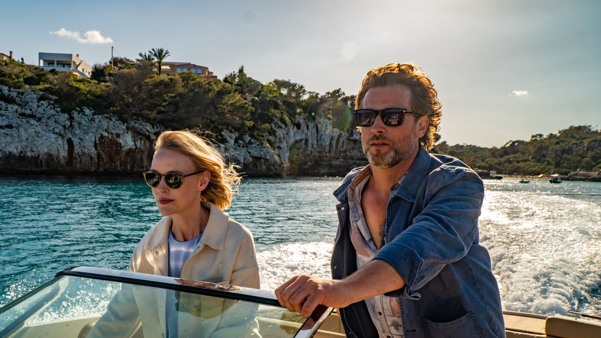 "The Mallorca Files - Nicht alles ist Gold was glänzt": Miranda (Elen Rhys) und Max (Julian Looman) befinden sich in einem Motorboot, das vor einer felsigen Küste entlang fährt. Miranda hält konzentriert das Steuer, während Max neben ihr steht und in die Ferne blickt.
