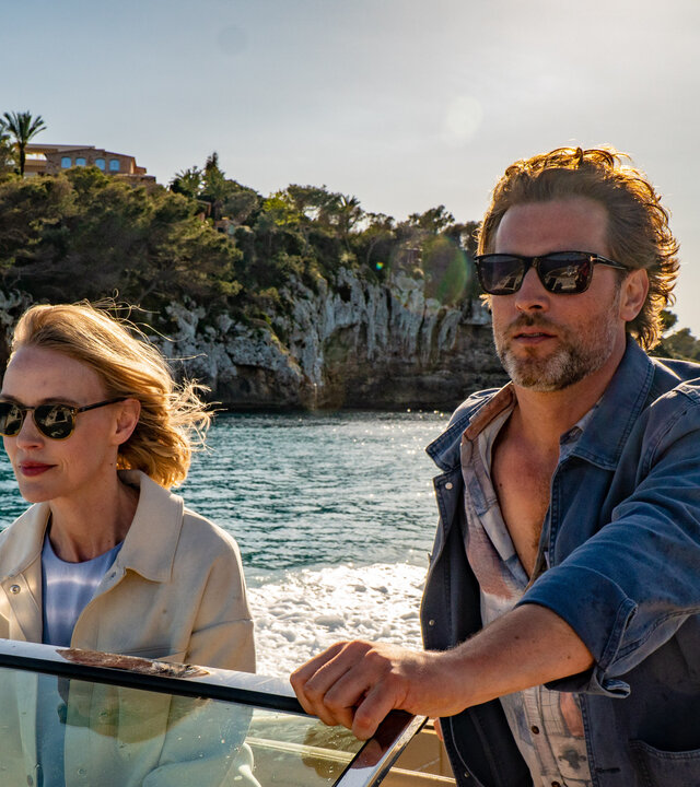 "The Mallorca Files - Nicht alles ist Gold was glänzt": Miranda (Elen Rhys) und Max (Julian Looman) befinden sich in einem Motorboot, das vor einer felsigen Küste entlang fährt. Miranda hält konzentriert das Steuer, während Max neben ihr steht und in die Ferne blickt.