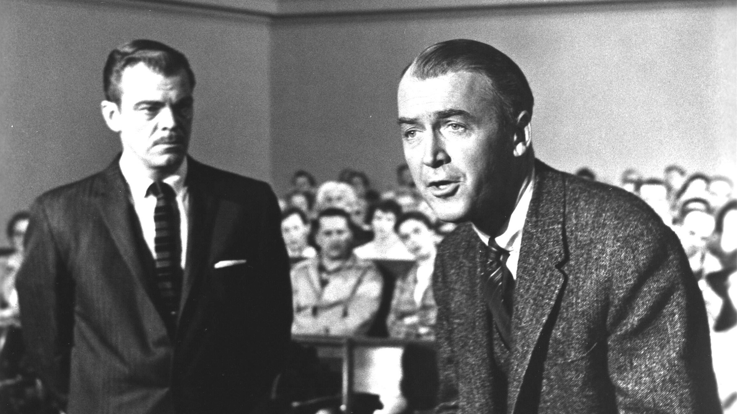 "Anatomie eines Mordes": Ein Gerichtssaal; vollbesetzte Zuschauerreihen unscharf im Hintergrund. Vorne stehend Paul Biegler (James Stewart), der mit geballten Fäusten in engagierter Haltung das Wort führt. Neben ihm, leicht versetzt nach hinten Mitch Lodwick (Brooks West), der ihn stumm und mit auf dem Rücken verschränkten Armen beobachtet.