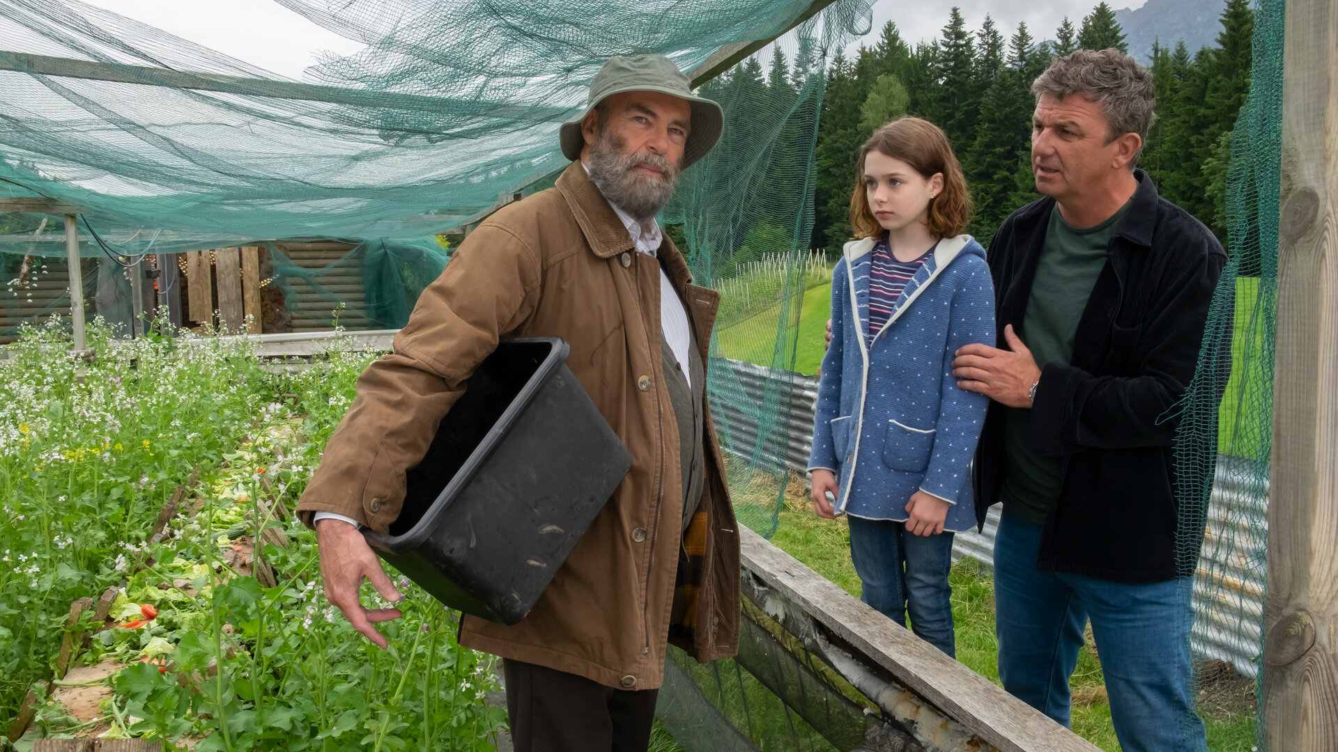 "Der Bergdoktor - Der Blick nach vorn": Im Garten: Martin Gruber (Hans Sigl) und Emmi Neumann (Lotta Herzog) blicken erwartungsvoll Anton Grainer (Peter Kremer) an. Dieser hat den Blick von beiden abgewandt und blickt nachdenklich in die Ferne. Er hält eine große Plastikwanne unter dem Arm und trägt einen Hut.