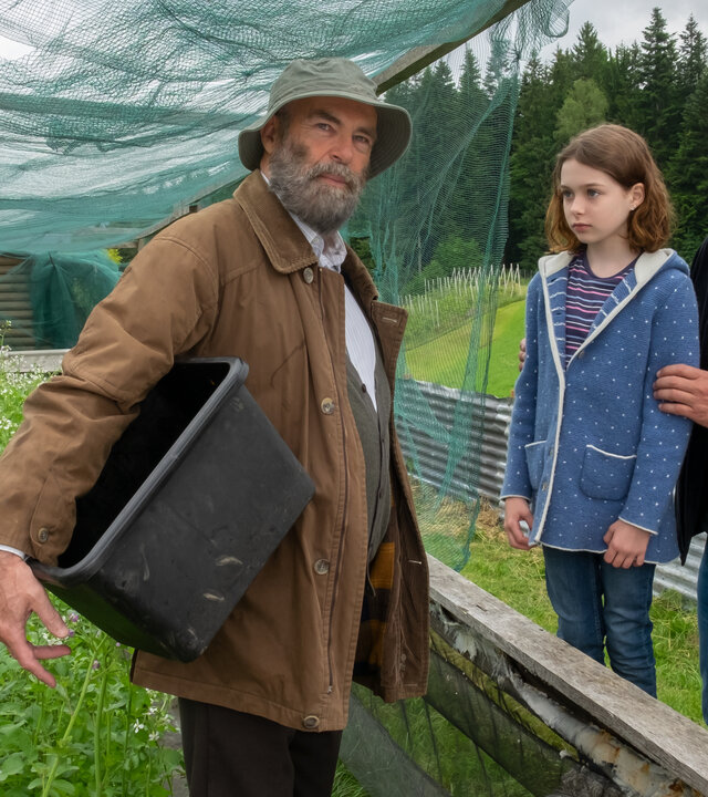"Der Bergdoktor - Der Blick nach vorn": Im Garten: Martin Gruber (Hans Sigl) und Emmi Neumann (Lotta Herzog) blicken erwartungsvoll Anton Grainer (Peter Kremer) an. Dieser hat den Blick von beiden abgewandt und blickt nachdenklich in die Ferne. Er hält eine große Plastikwanne unter dem Arm und trägt einen Hut.