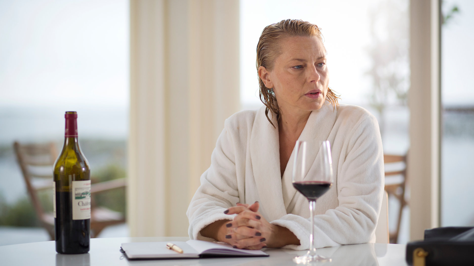 "Springflut - Das Vermächtnis": Linn Magnusson (Helena Bergström) sitzt im weißen Bademantel mit nassen, zurückgekämmten Haaren am Esstisch. Vor ihr sind ein Rotweinglas, eine Flasche Rotwein und ein aufgeschlagenes Buch mit Kugelschreiber zu sehen.