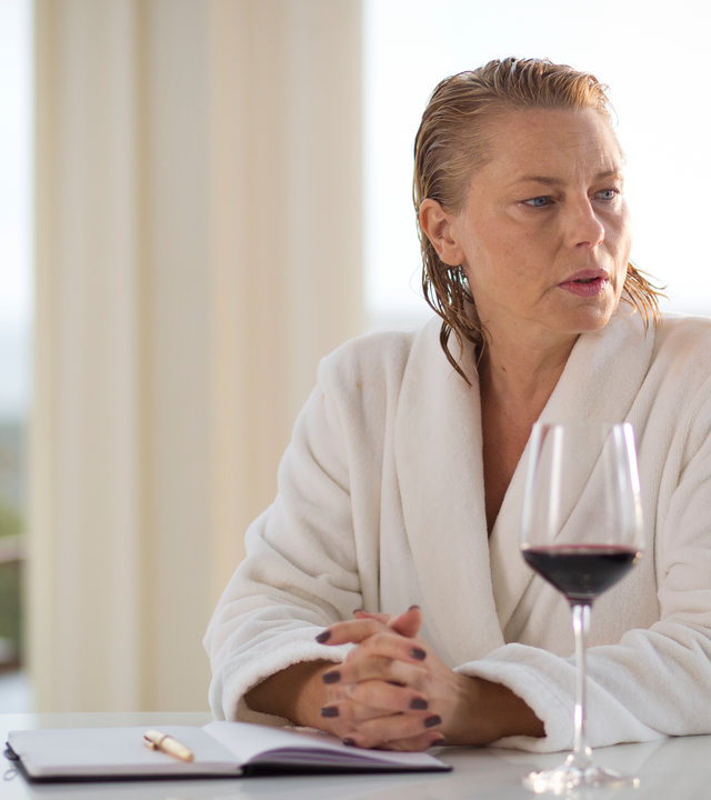 "Springflut - Das Vermächtnis": Linn Magnusson (Helena Bergström) sitzt im weißen Bademantel mit nassen, zurückgekämmten Haaren am Esstisch. Vor ihr sind ein Rotweinglas, eine Flasche Rotwein und ein aufgeschlagenes Buch mit Kugelschreiber zu sehen.