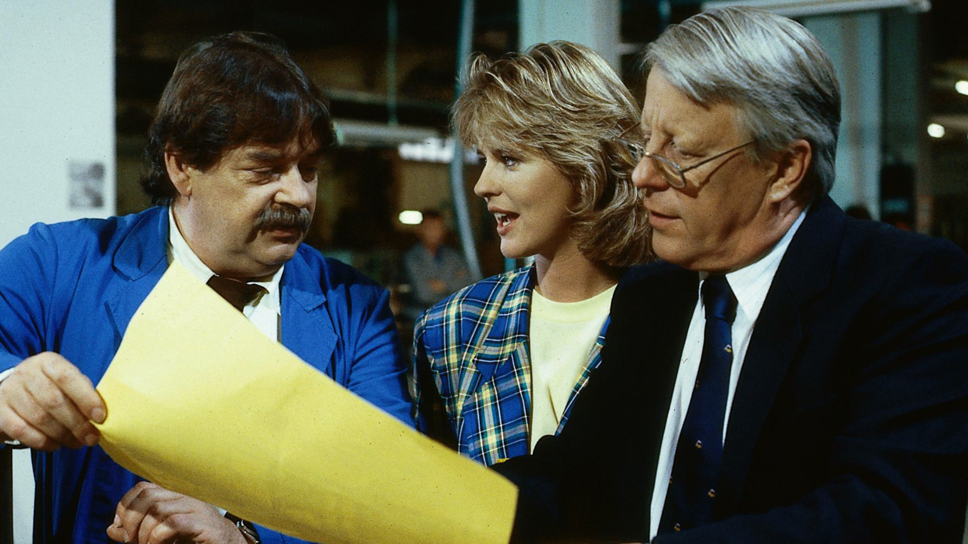 "Die Wicherts von nebenan: Ernste Lage": Eberhard (Stephan Orlac), Werkmeister Heinz (Andreas Mannkopff) und Uschi (Anja Schüte) stehen nebeneinander und blicken auf einen Bogen Papier, den die Männer in der Hand halten.