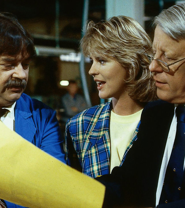 "Die Wicherts von nebenan: Ernste Lage": Eberhard (Stephan Orlac), Werkmeister Heinz (Andreas Mannkopff) und Uschi (Anja Schüte) stehen nebeneinander und blicken auf einen Bogen Papier, den die Männer in der Hand halten.