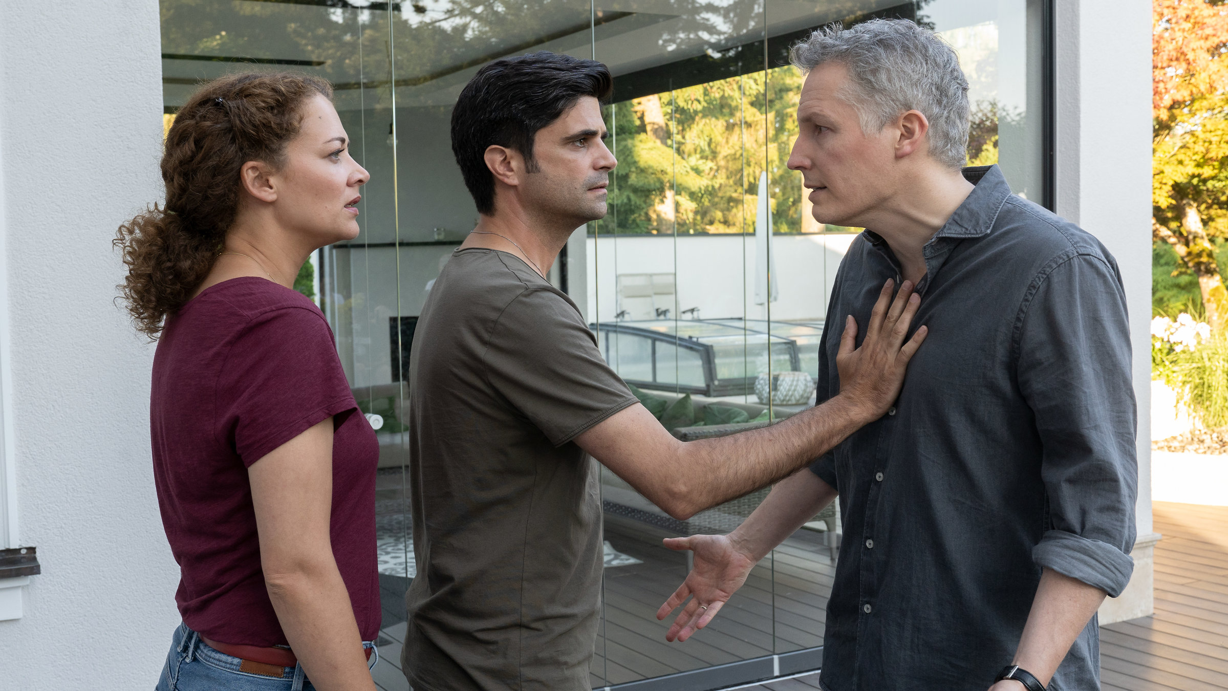 "Tonio & Julia - Der perfekte Mann": Auf einer Terrasse vor einer Glaspanoramatür, die einen Blick in einen Wohnraum und den seitlich davon liegenden Pool eröffnet, stehen Julia Schindel (Oona Devi Liebich), Tonio Niederegger (Maximilian Grill) und Joachim Stegmeyer (Max Urlacher). Stegmeyer scheint aufgebracht. Daher hält Tonio ihn mit der Hand auf dessen Brust zurück, während Julia mit ihm spricht.