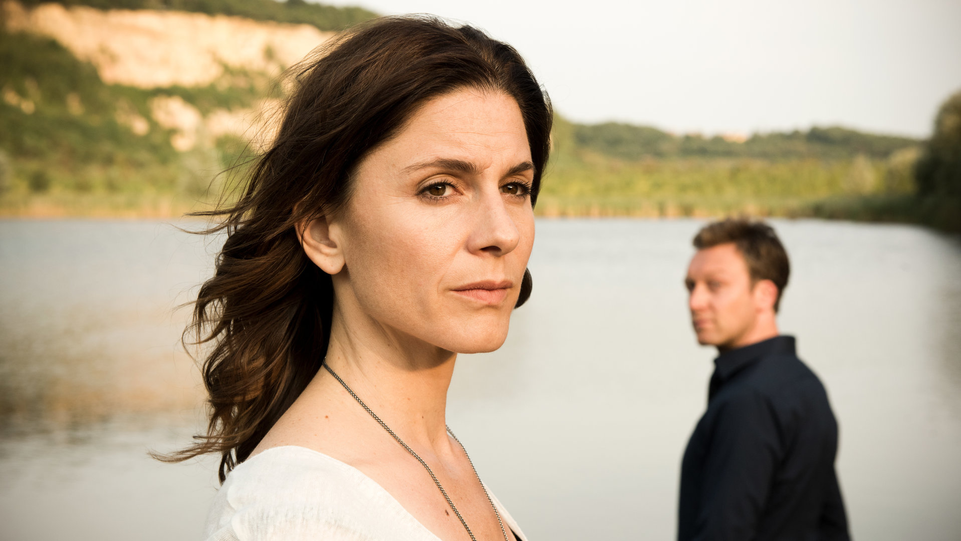 "In Wahrheit - Still ruht der See": Judith Mohn (Christina Hecke) blickt nachdenklich aus dem Bild. Hinter ihr steht Freddy Breyer (Robin Sondermann) und schaut zu Judith hin. Im Hintergrund ist ein See zu sehen.